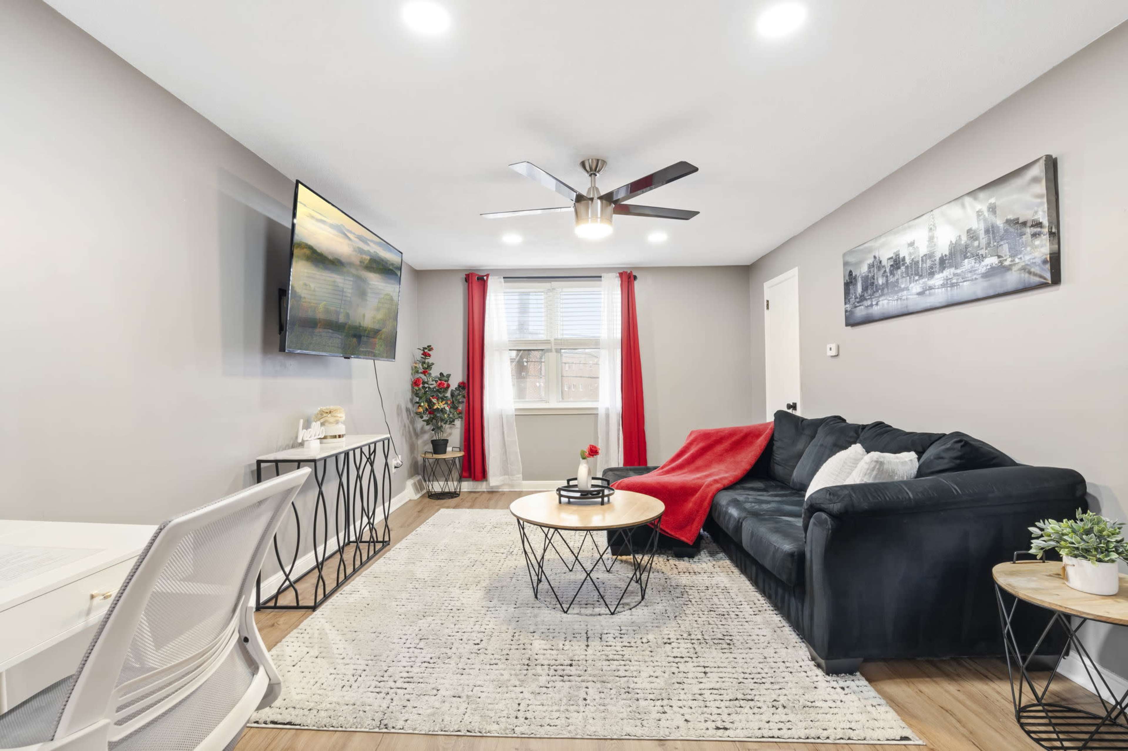 A modern living room features a black sofa, a circular coffee table, a mounted TV, and decorative elements including a plant and red curtains.
