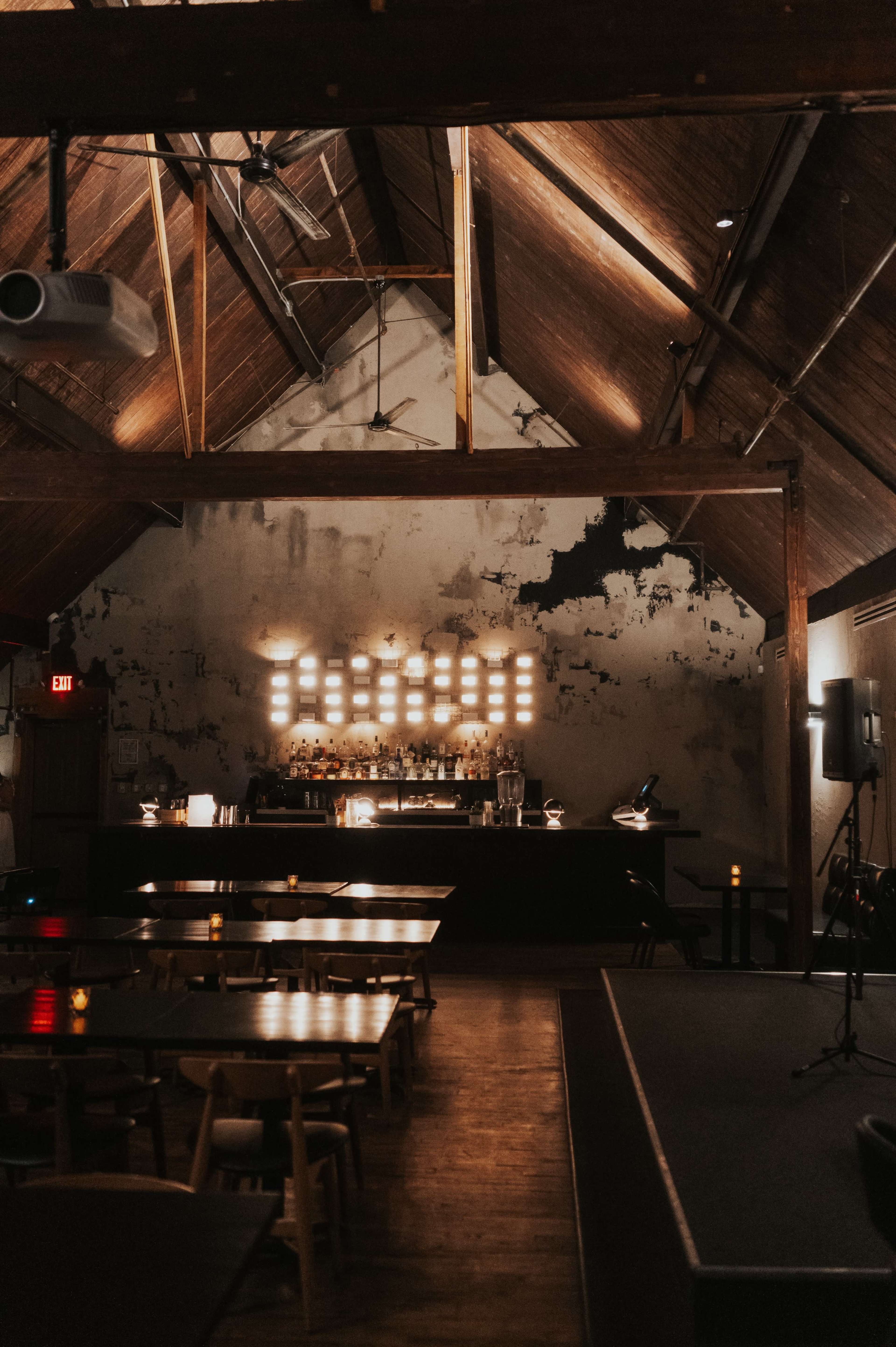 The image shows a dimly lit restaurant interior featuring wooden tables and a bar with a backlit display against a rustic wall.