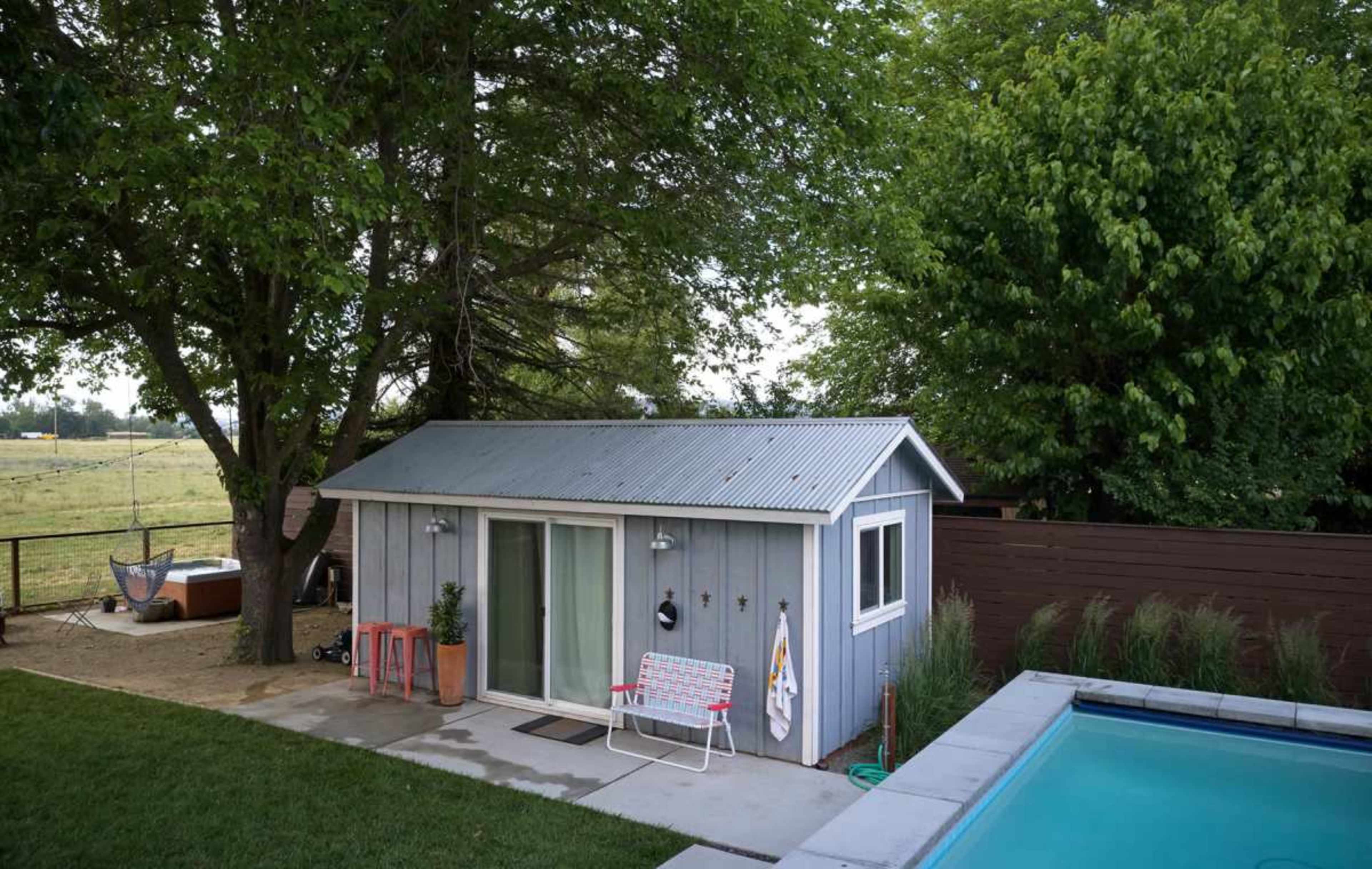 A small gray backyard shed with a metal roof is situated next to a swimming pool, surrounded by trees and grassy areas.
