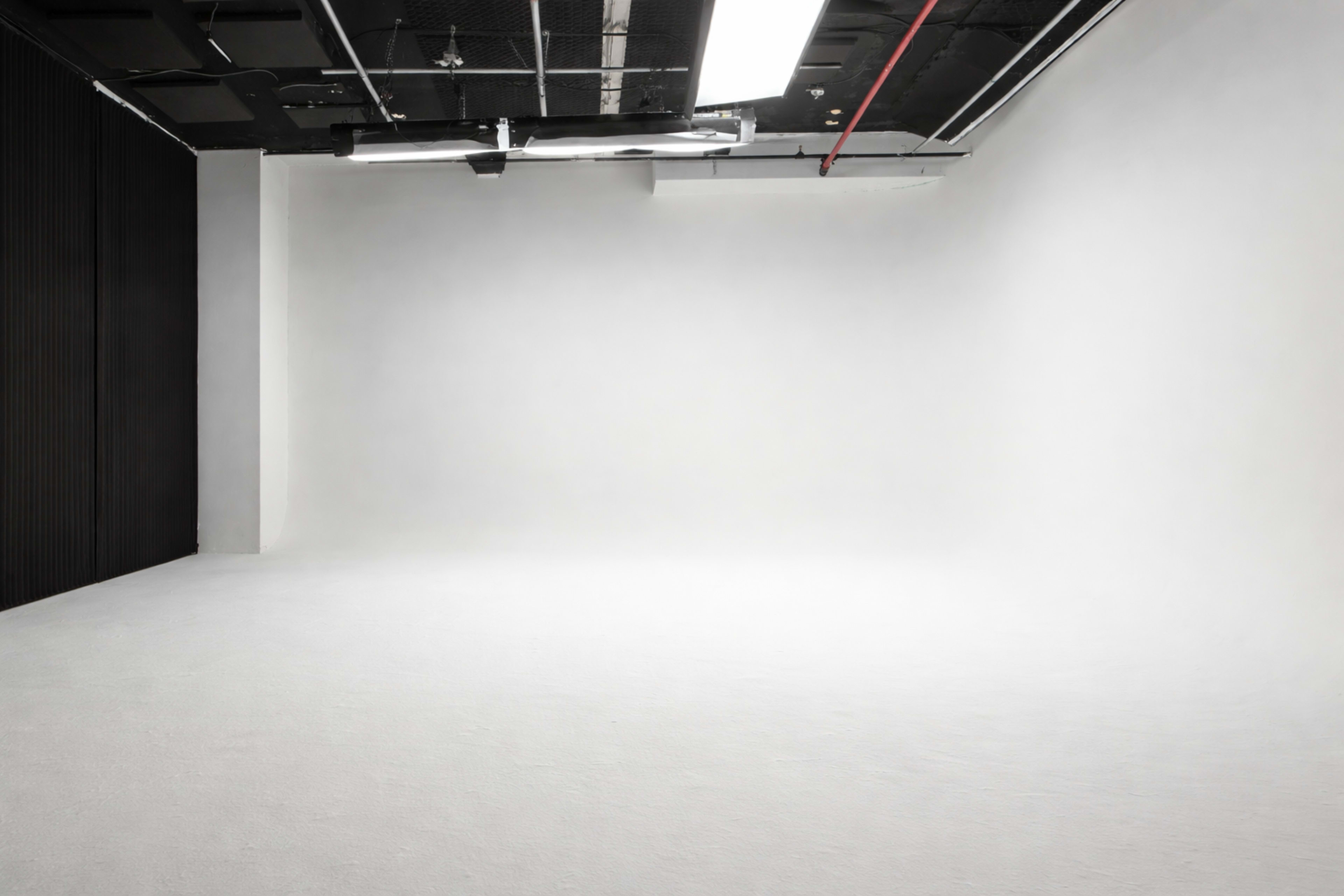 The image shows an empty, minimalist studio space with white walls and a smooth floor, illuminated by overhead lights.