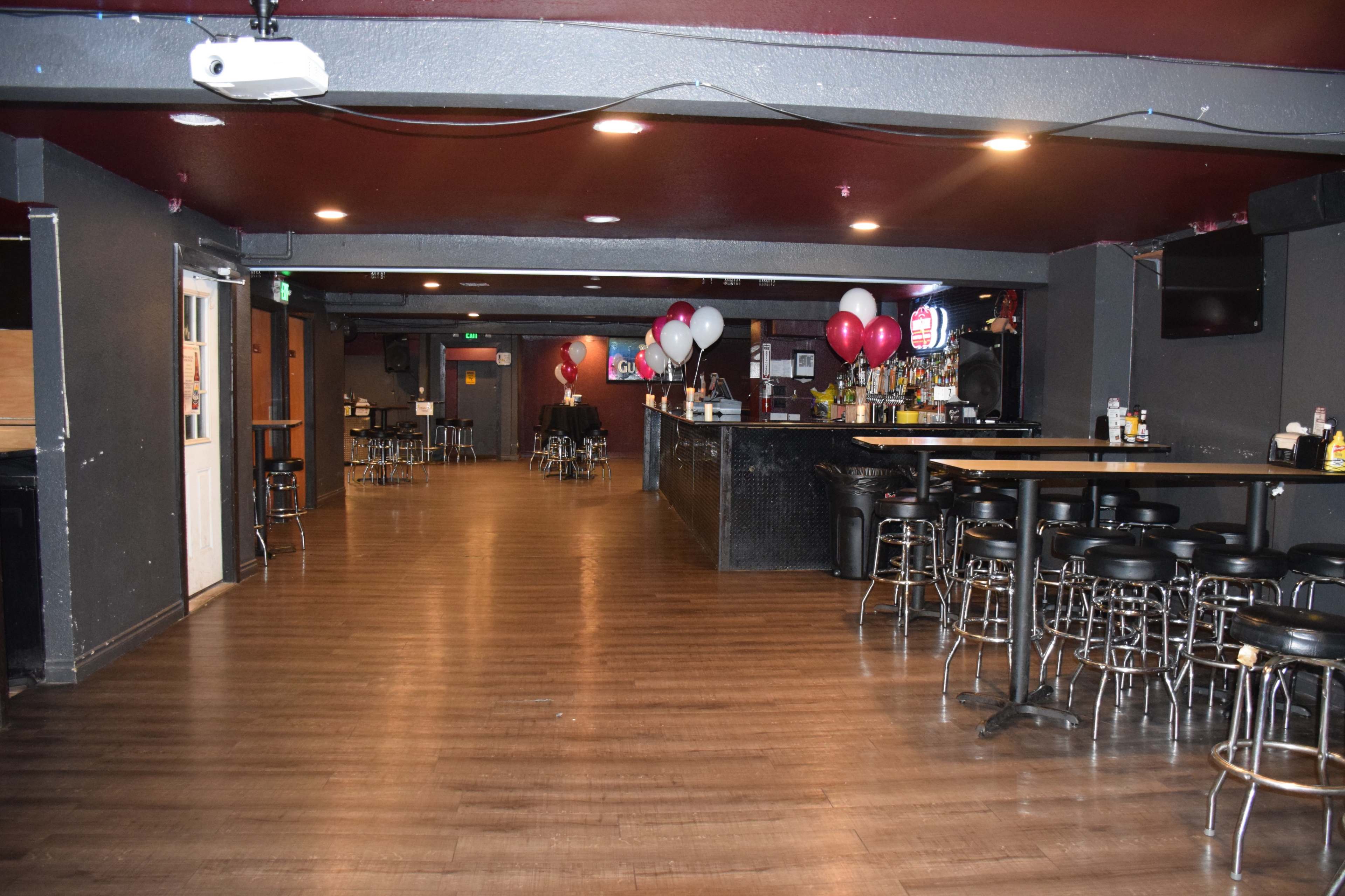 A spacious bar area with black walls, wooden flooring, several tables, chairs, and balloons arranged at one end.