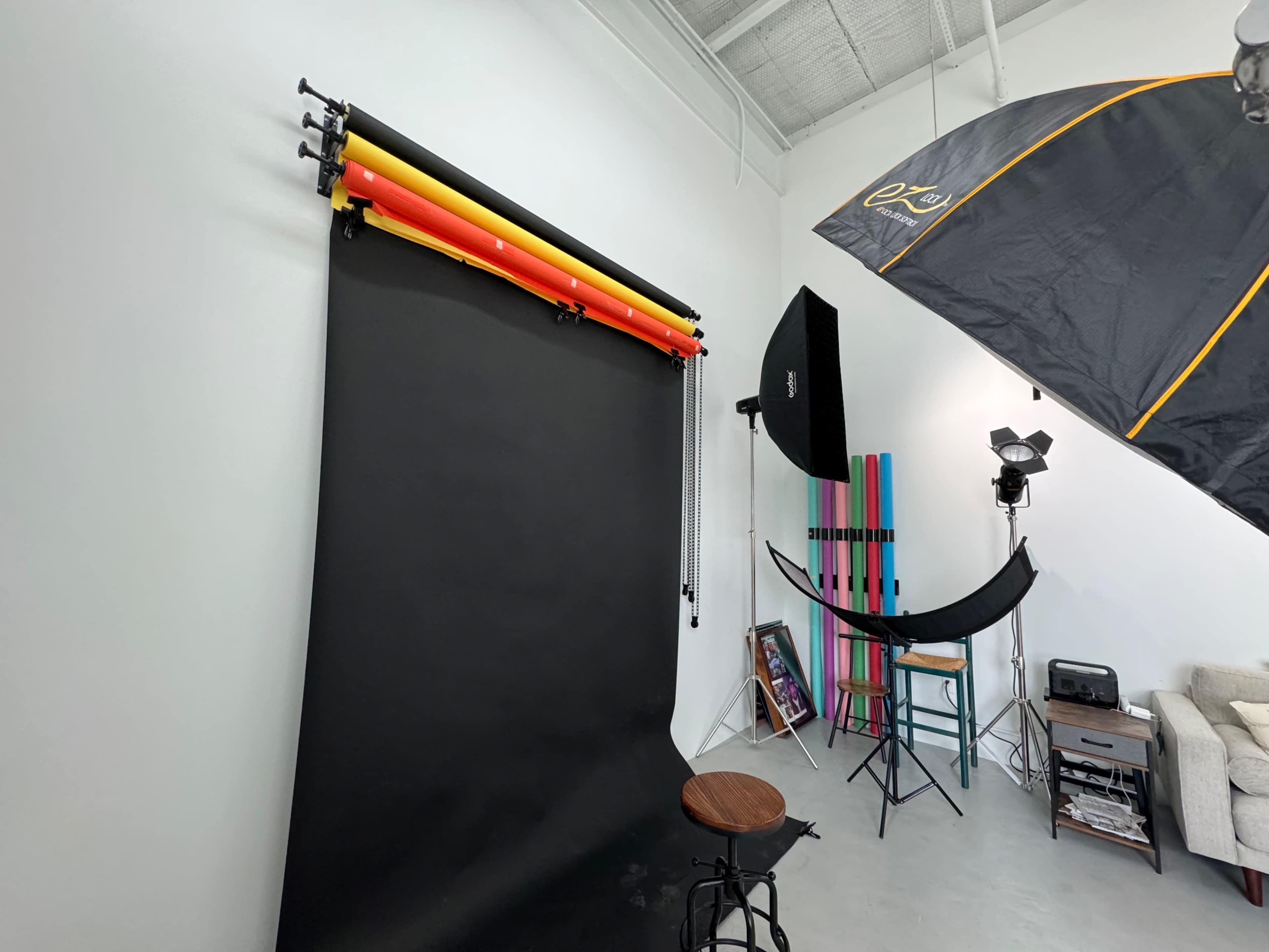 The image shows a photography studio with a black backdrop, colorful backdrop rolls, various lighting equipment, and a seating area.