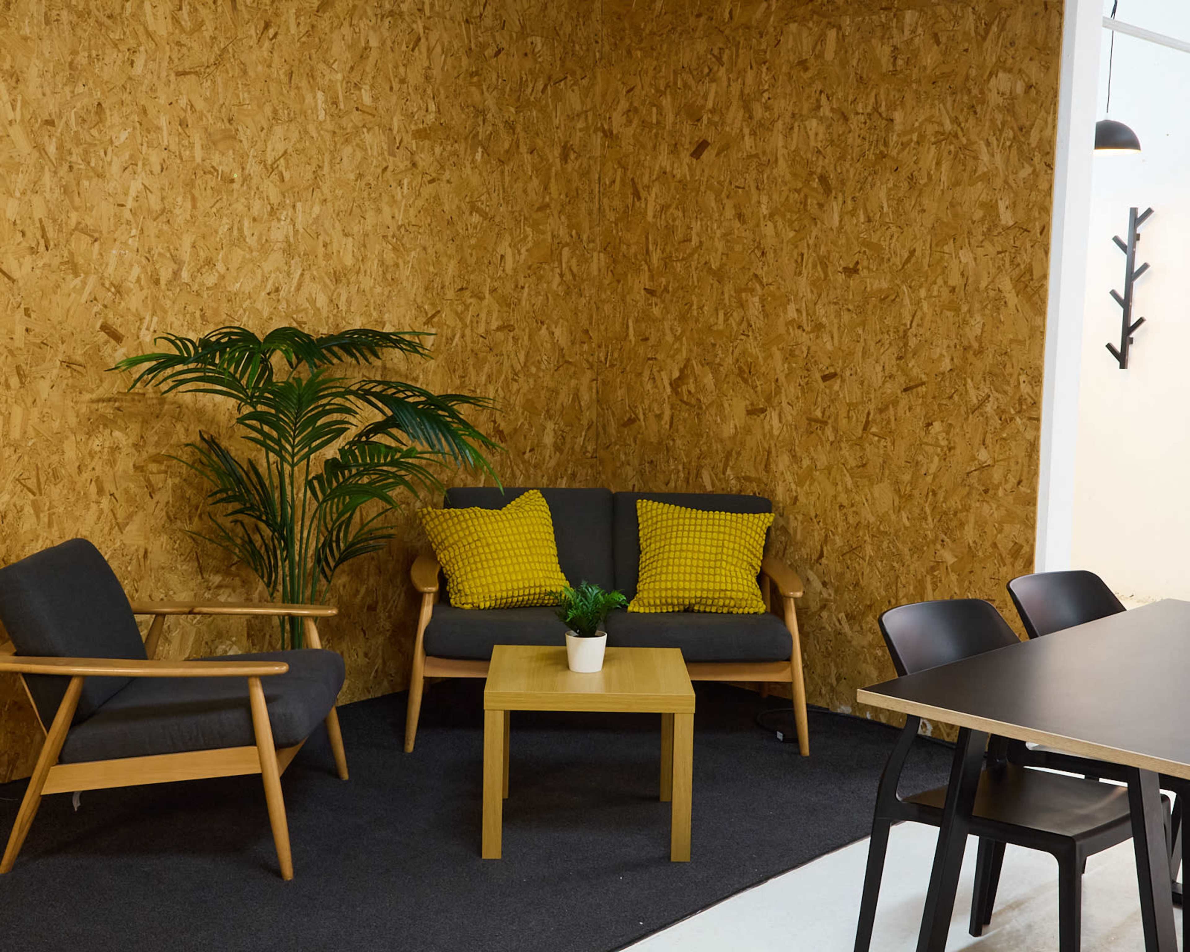 A cozy seating area features a gray couch with yellow pillows, a small wooden coffee table, a potted plant, and a black dining table against an OSB wood wall.