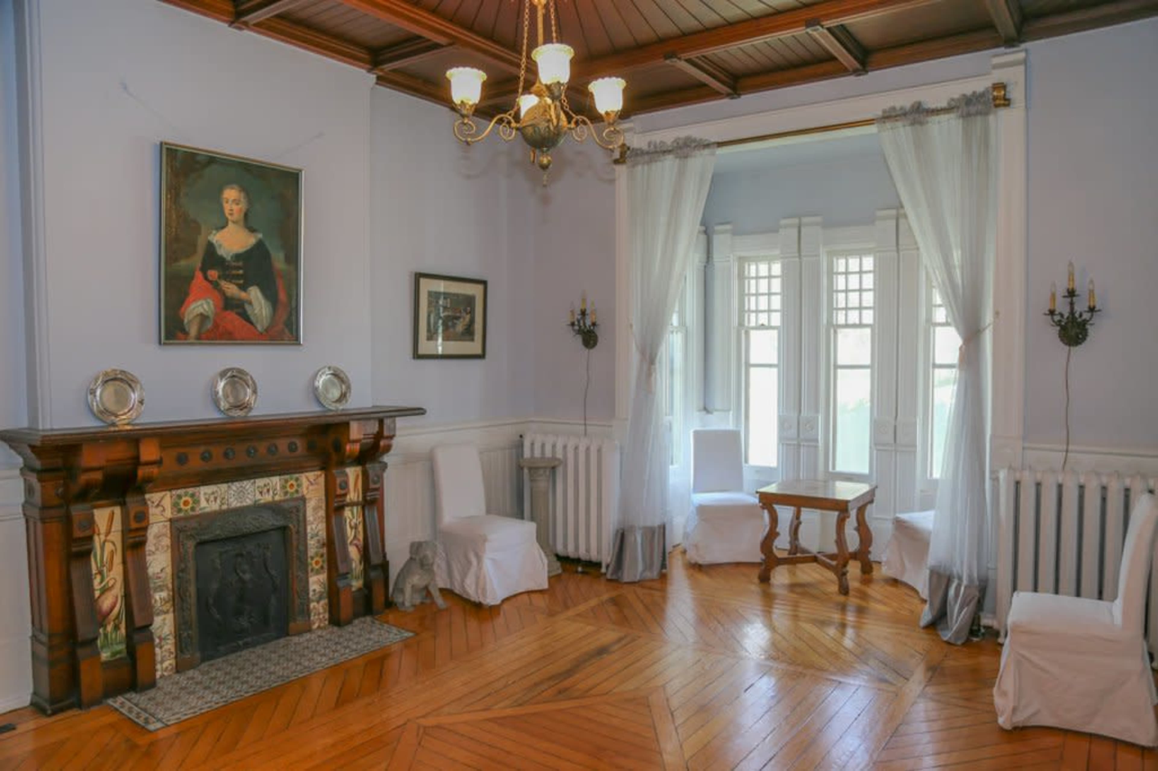 The room features a wooden fireplace adorned with decorative tiles, surrounded by chairs covered in white cloth, and large windows framed by sheer curtains.