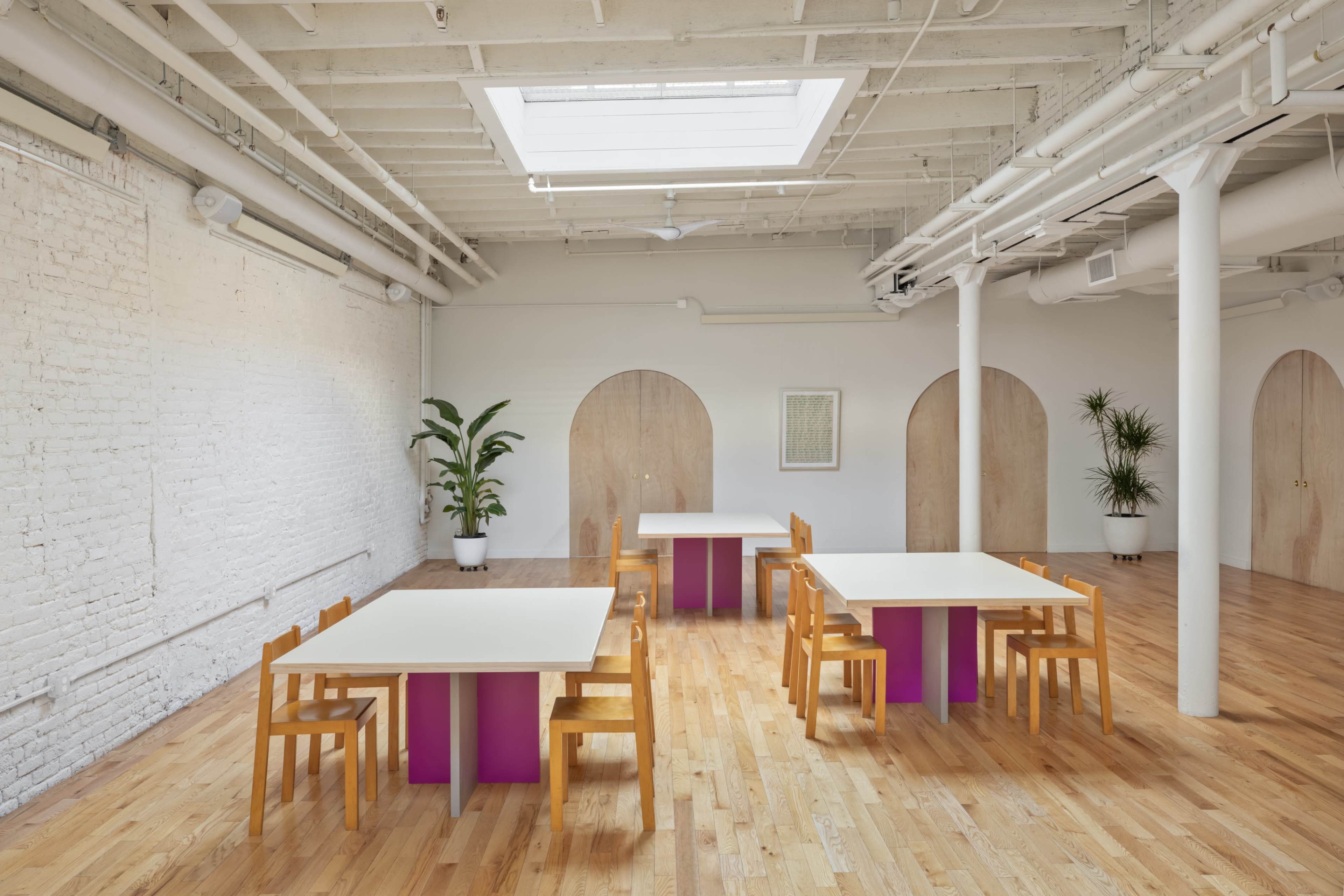 A spacious room with wooden floors, featuring several tables with light-colored surfaces and wooden chairs, complemented by potted plants and a skylight.