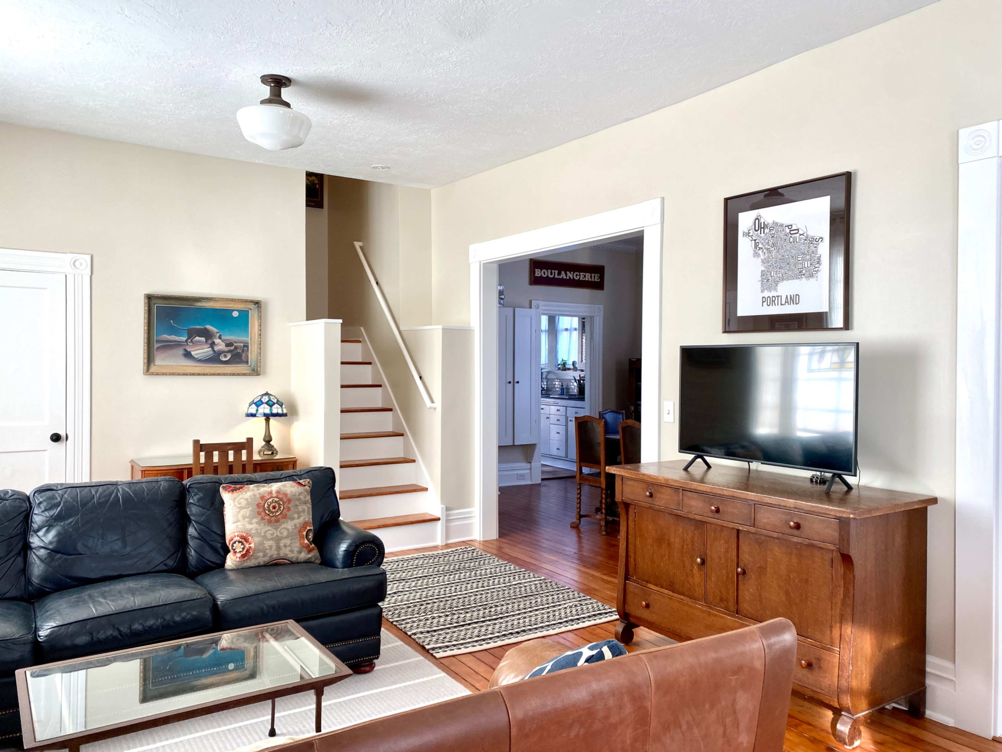 A living room features a dark leather sofa, a glass-top coffee table, a wooden sideboard with a television, and a staircase leading to the upper level.
