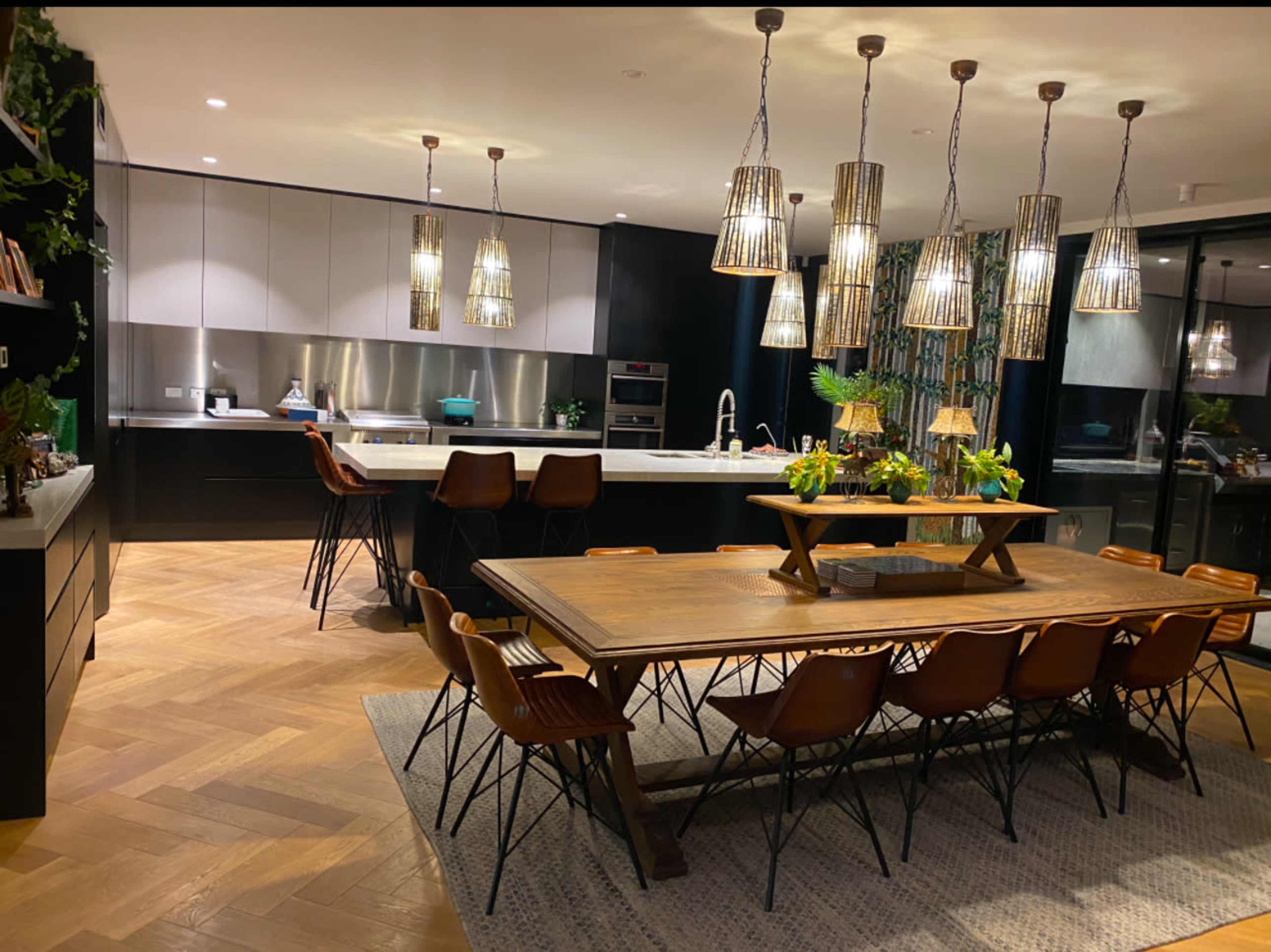 The image shows a modern kitchen and dining area featuring a long wooden dining table surrounded by brown chairs, illuminated by pendant lights.