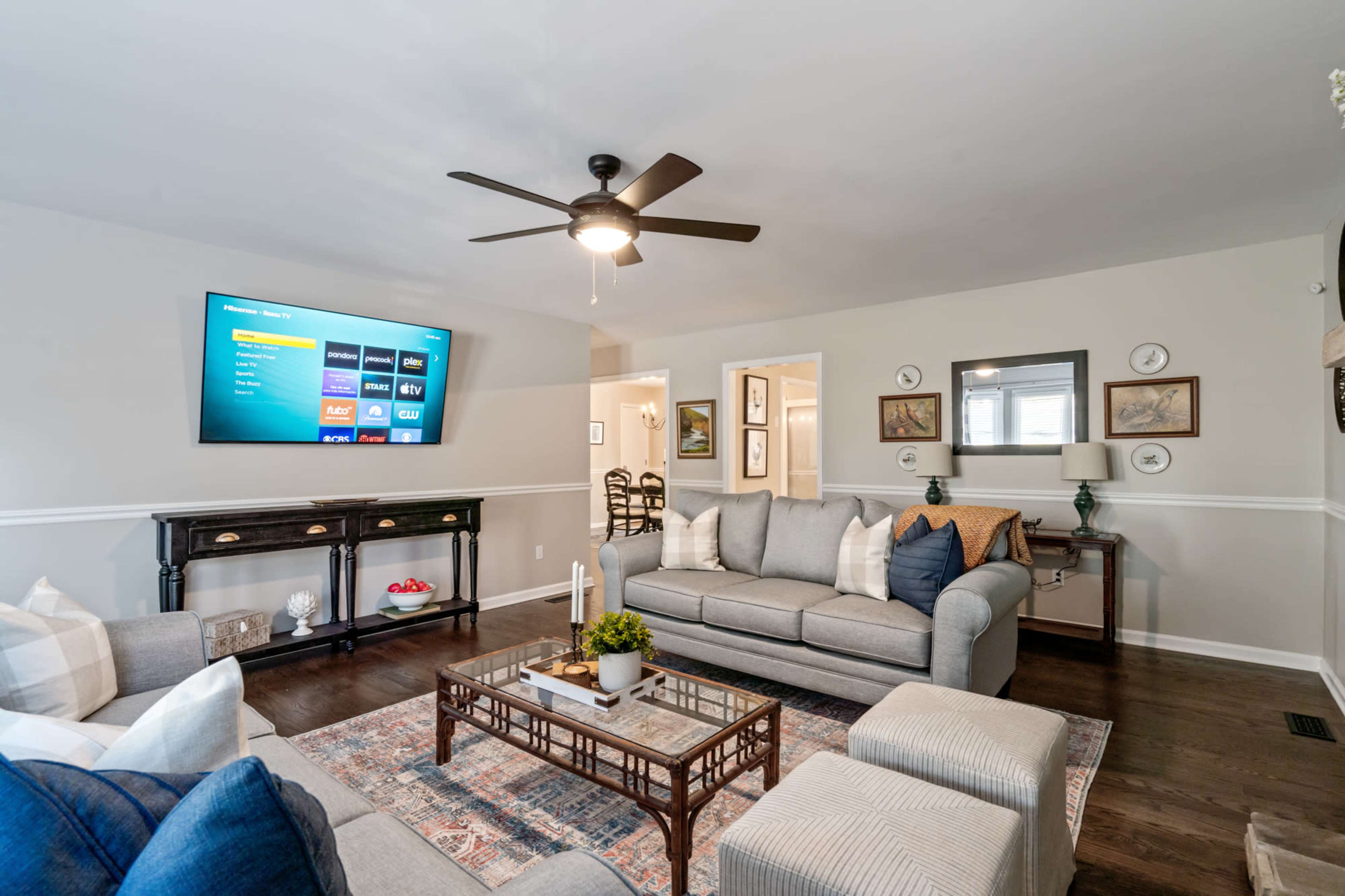 A cozy living room with a large flat-screen TV mounted on the wall, a gray sofa, and a coffee table on a patterned rug.