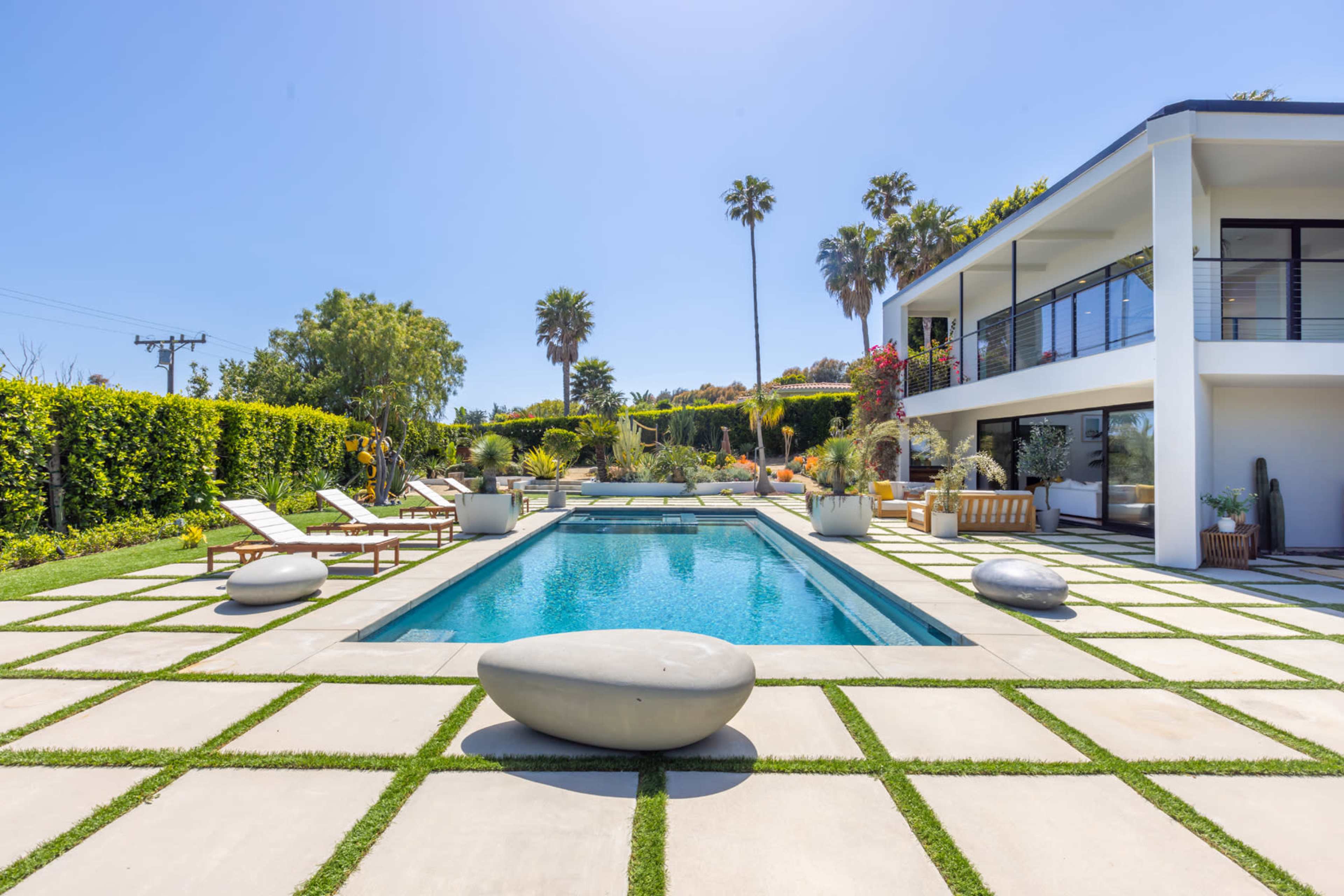A modern house with a rectangular swimming pool is surrounded by neatly arranged stone pavers and landscaped greenery.