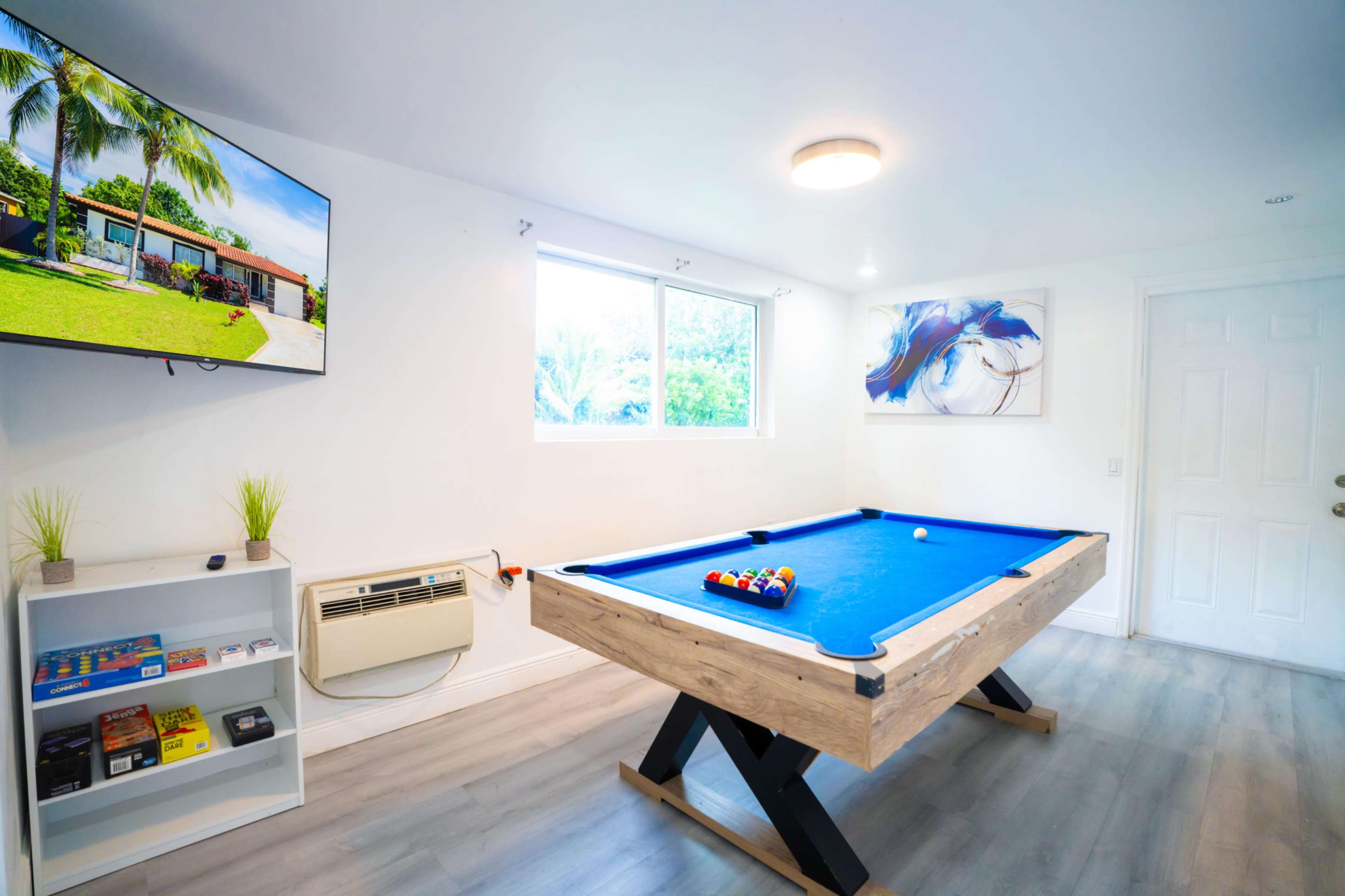 The image shows a bright game room featuring a blue pool table, a wall-mounted television, and a white shelf with various games.