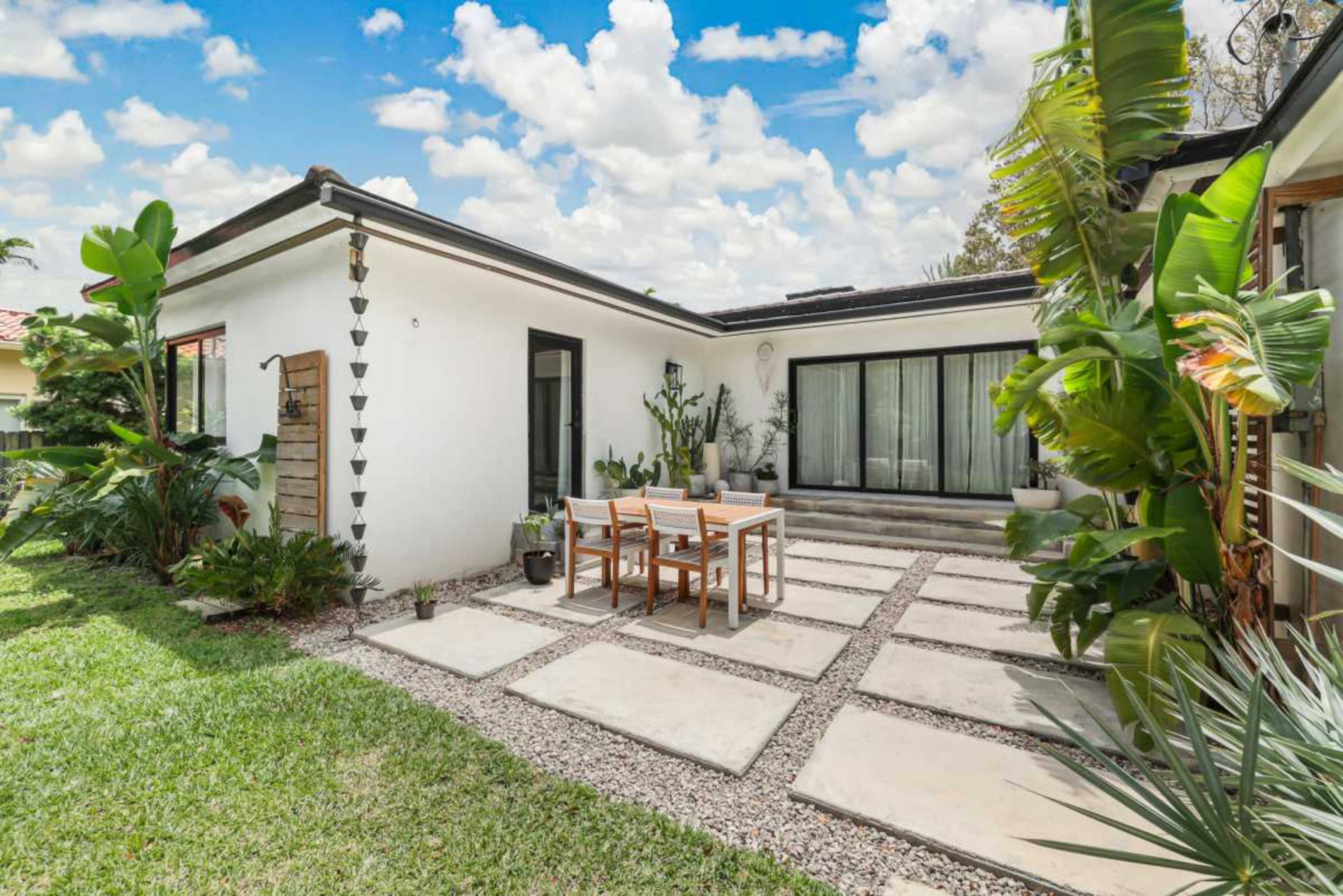 The image shows a landscaped outdoor area with a concrete patio, a wooden dining table, and lush greenery surrounding a modern home.