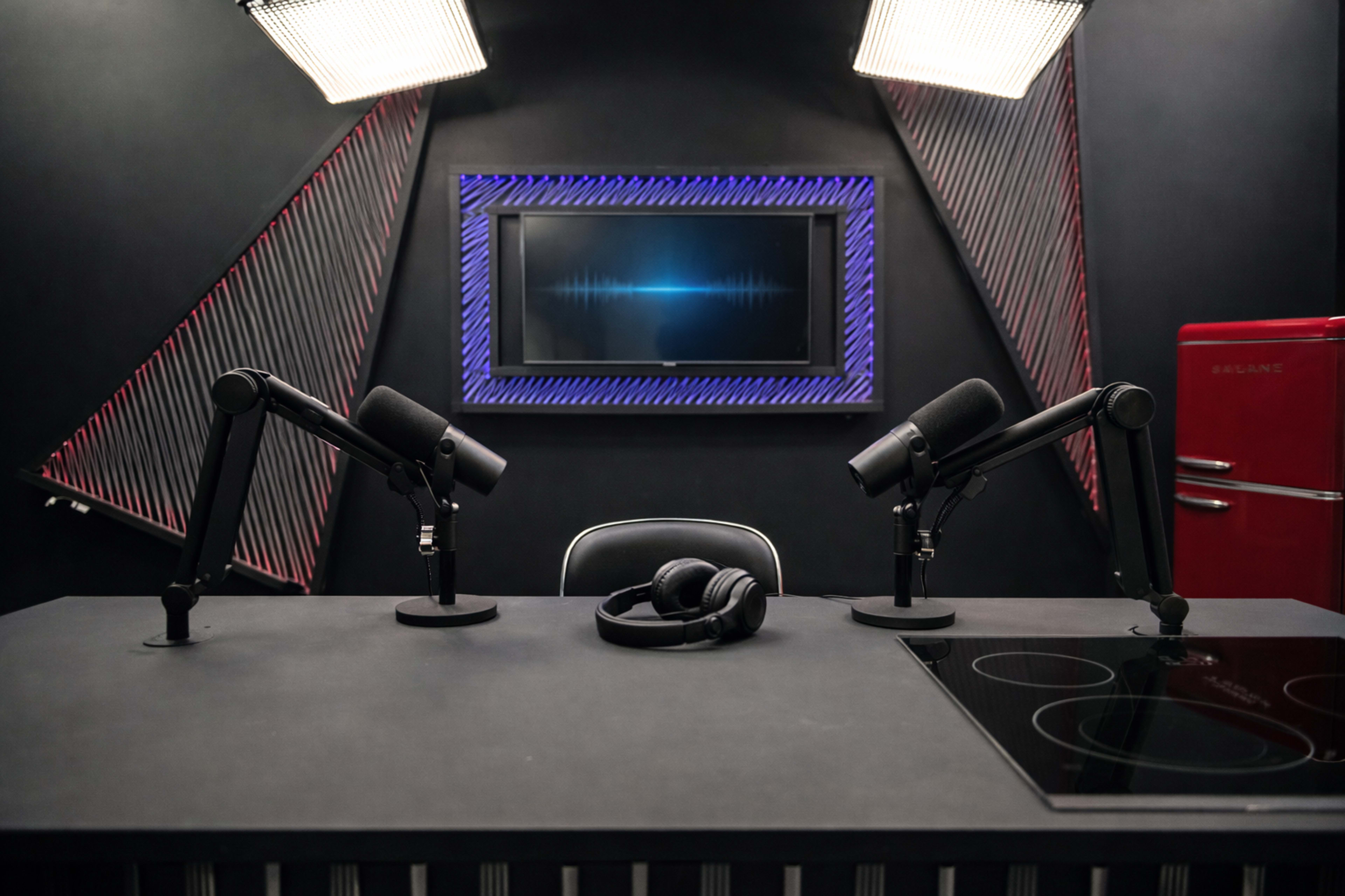 The image shows a modern podcast studio featuring a black table with two microphone stands, headphones, and a screen displaying a sound waveform against a dark backdrop.