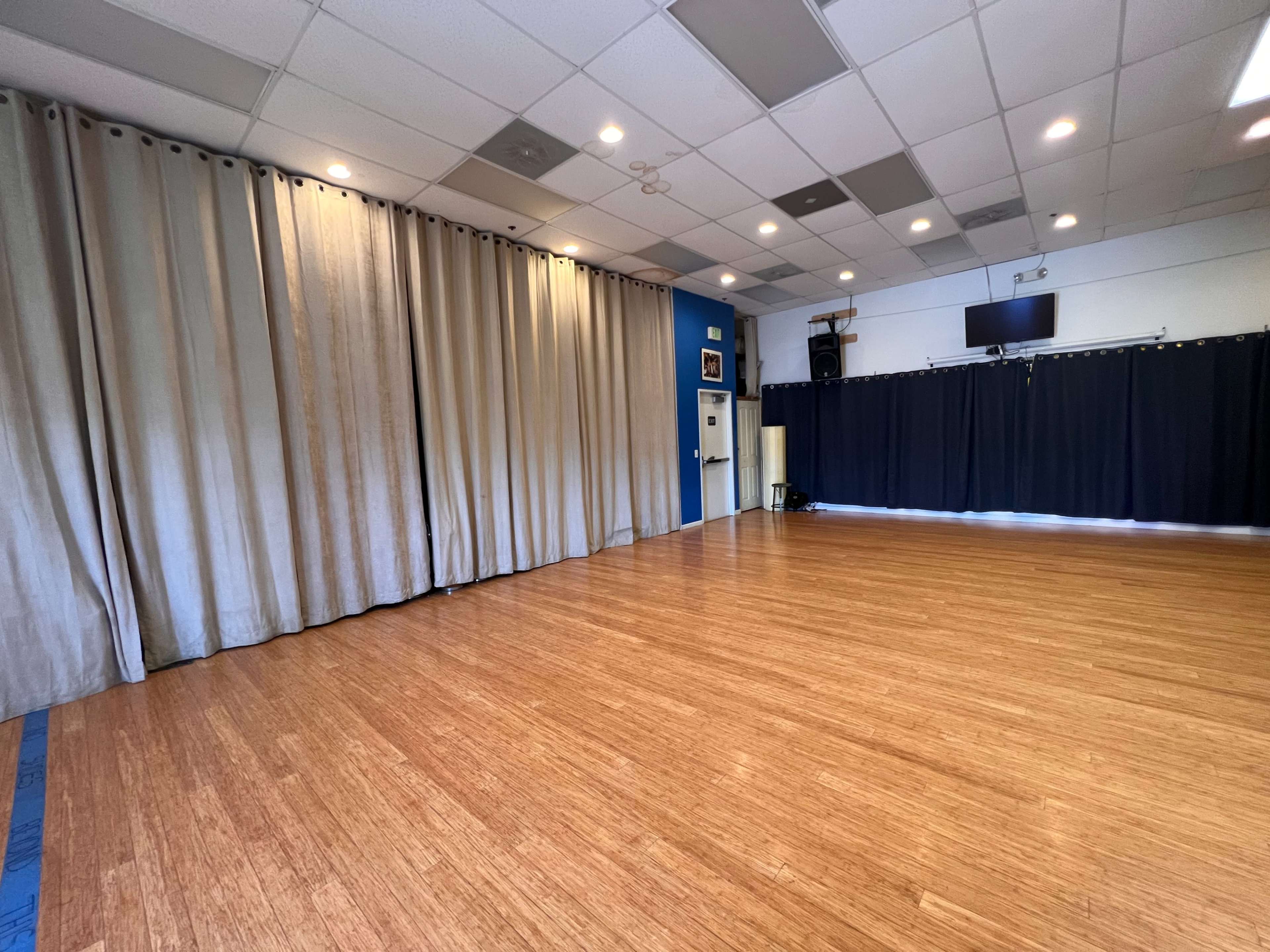 A spacious room with a wooden floor, dark curtains on one side, and beige curtains on the opposite side, along with wall-mounted equipment and lights.