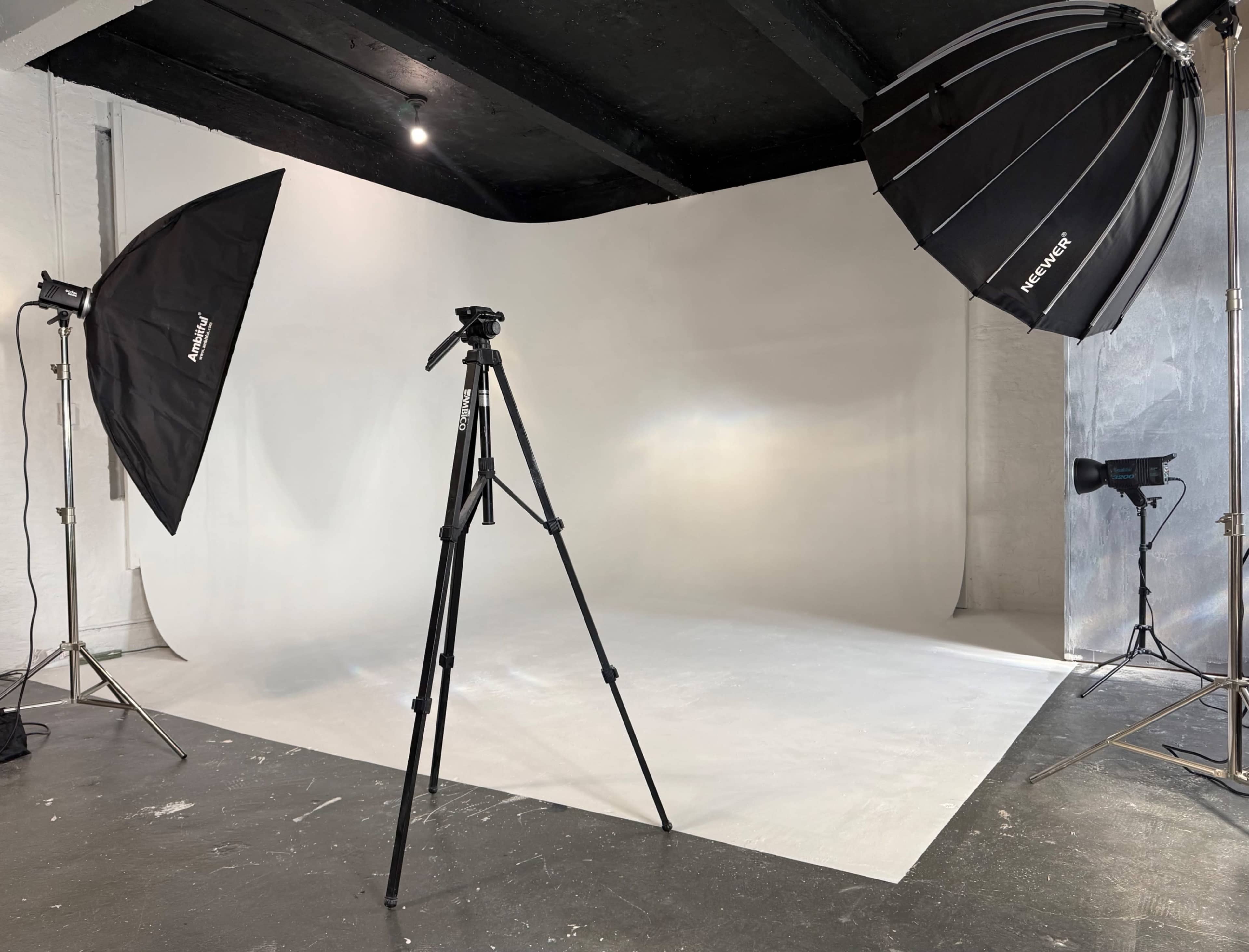 The image shows a photography studio with a plain backdrop, lighting equipment, and a tripod set up for a photo shoot.
