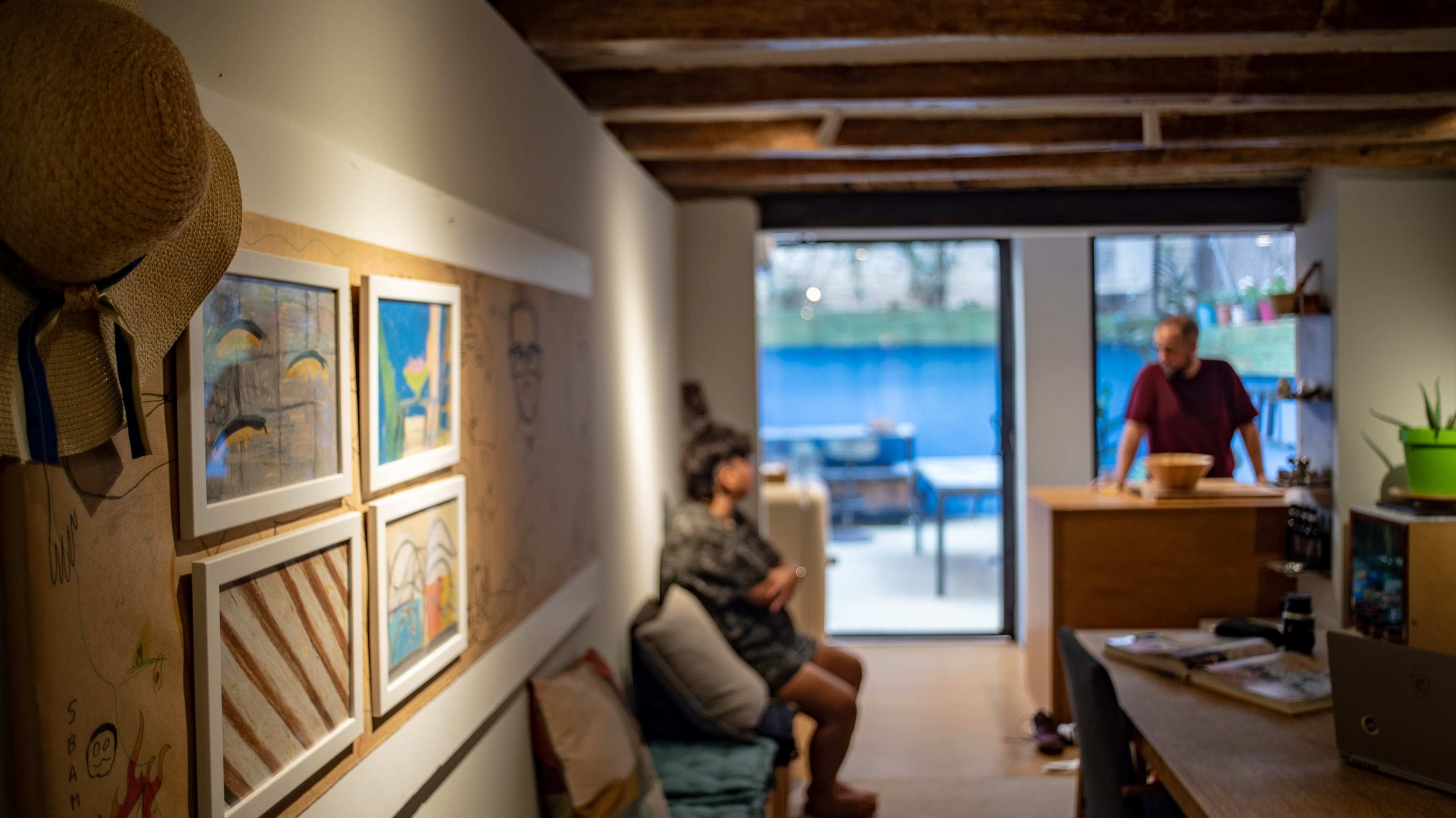 A cozy interior with a gallery of framed artwork on one wall, a person seated on a couch, and another individual standing by a wooden counter in a brightly lit space.