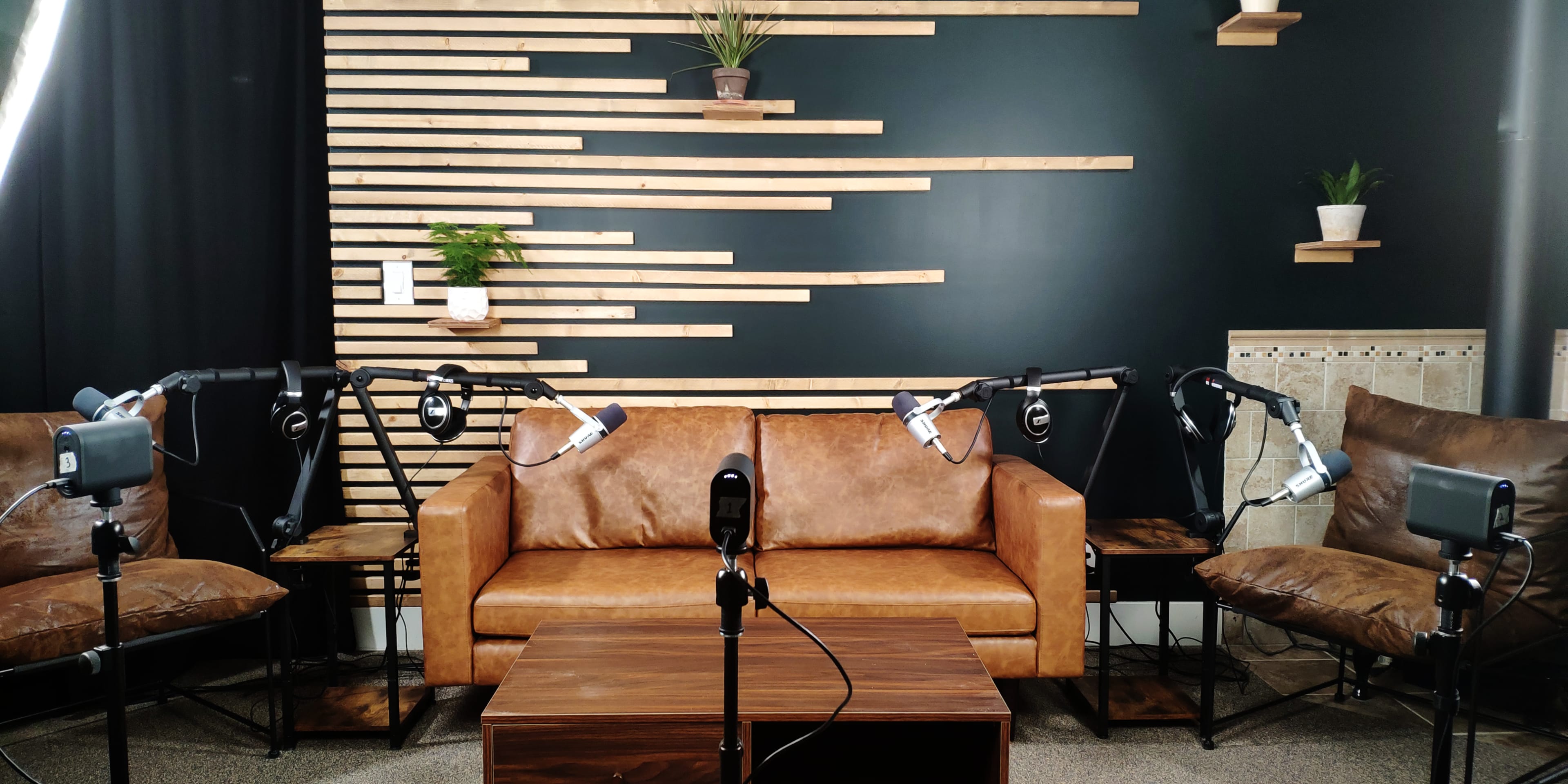 A modern seating area with a brown leather sofa, two armchairs, and several microphones positioned on stands, set against a wooden slat wall with plants.