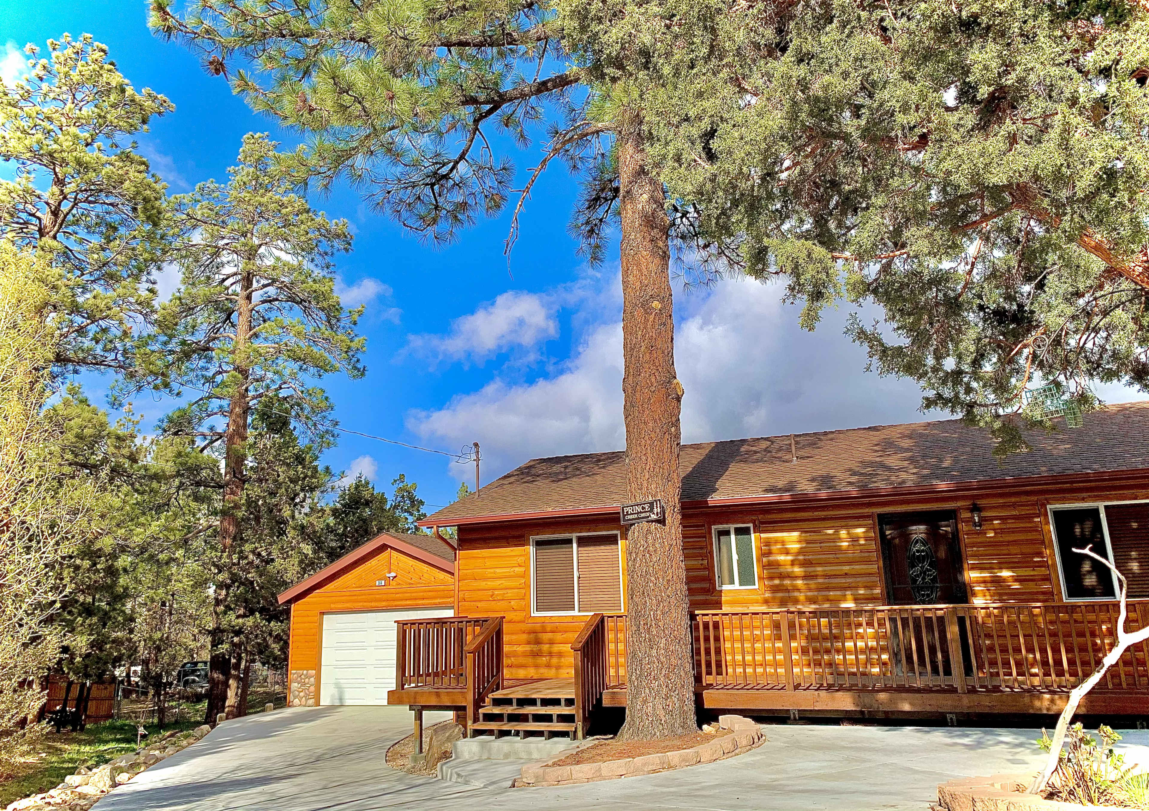 A brown wooden cabin with a porch and a garage is surrounded by tall green trees under a partly cloudy sky.