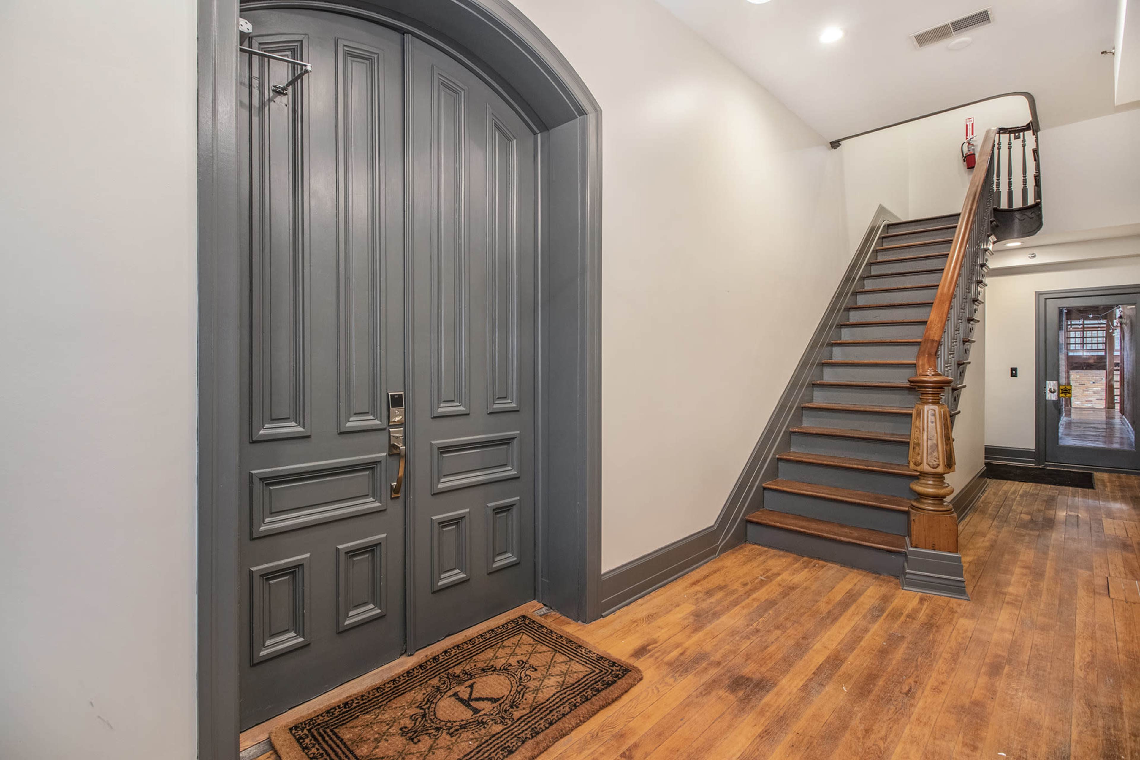 A hallway entrance with a large gray door, a wooden staircase leading to the upper floor, and a welcome mat on the floor.
