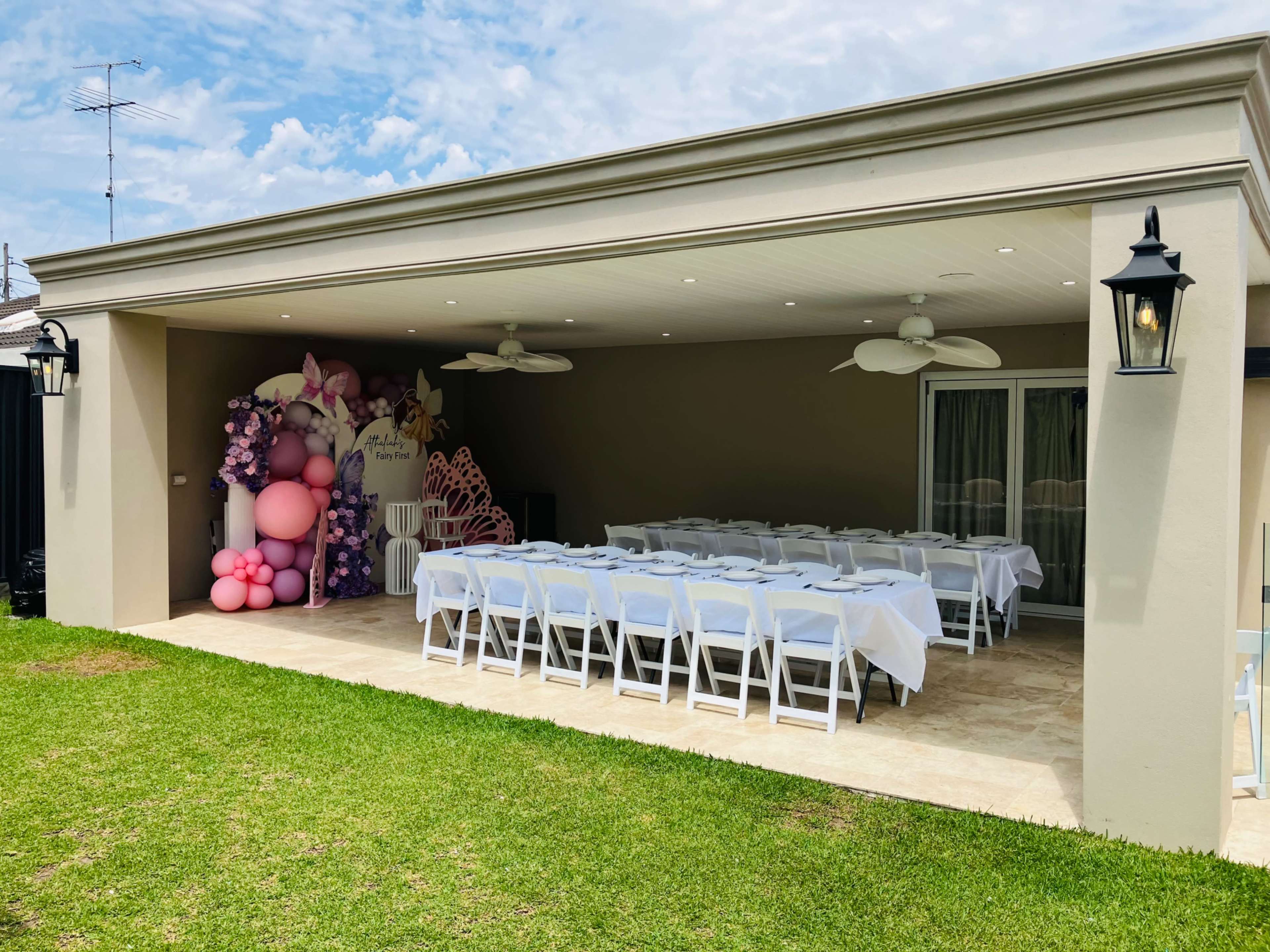 The image shows a covered outdoor space with white tables and chairs arranged for an event, featuring a floral backdrop and decorations in shades of pink.
