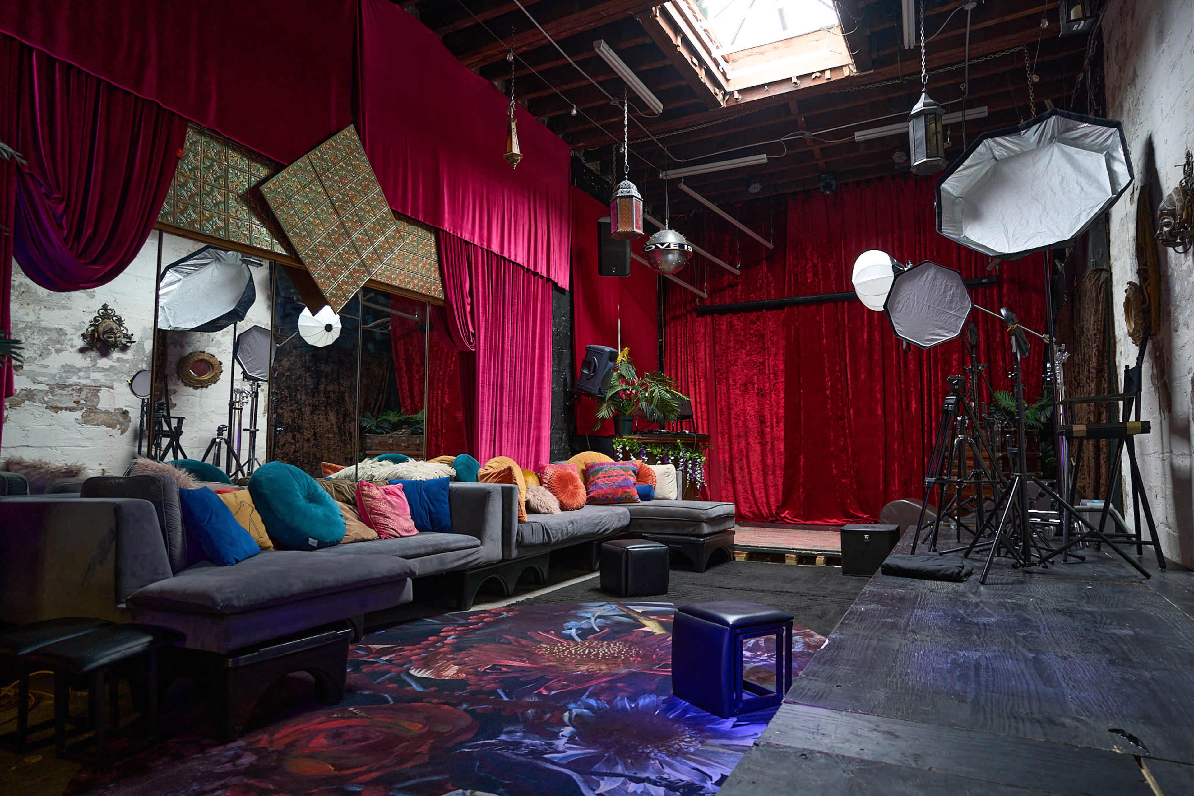 The image features a spacious photography studio with colorful cushions on a gray sofa, vibrant red curtains, and various lighting equipment set up for a shoot.