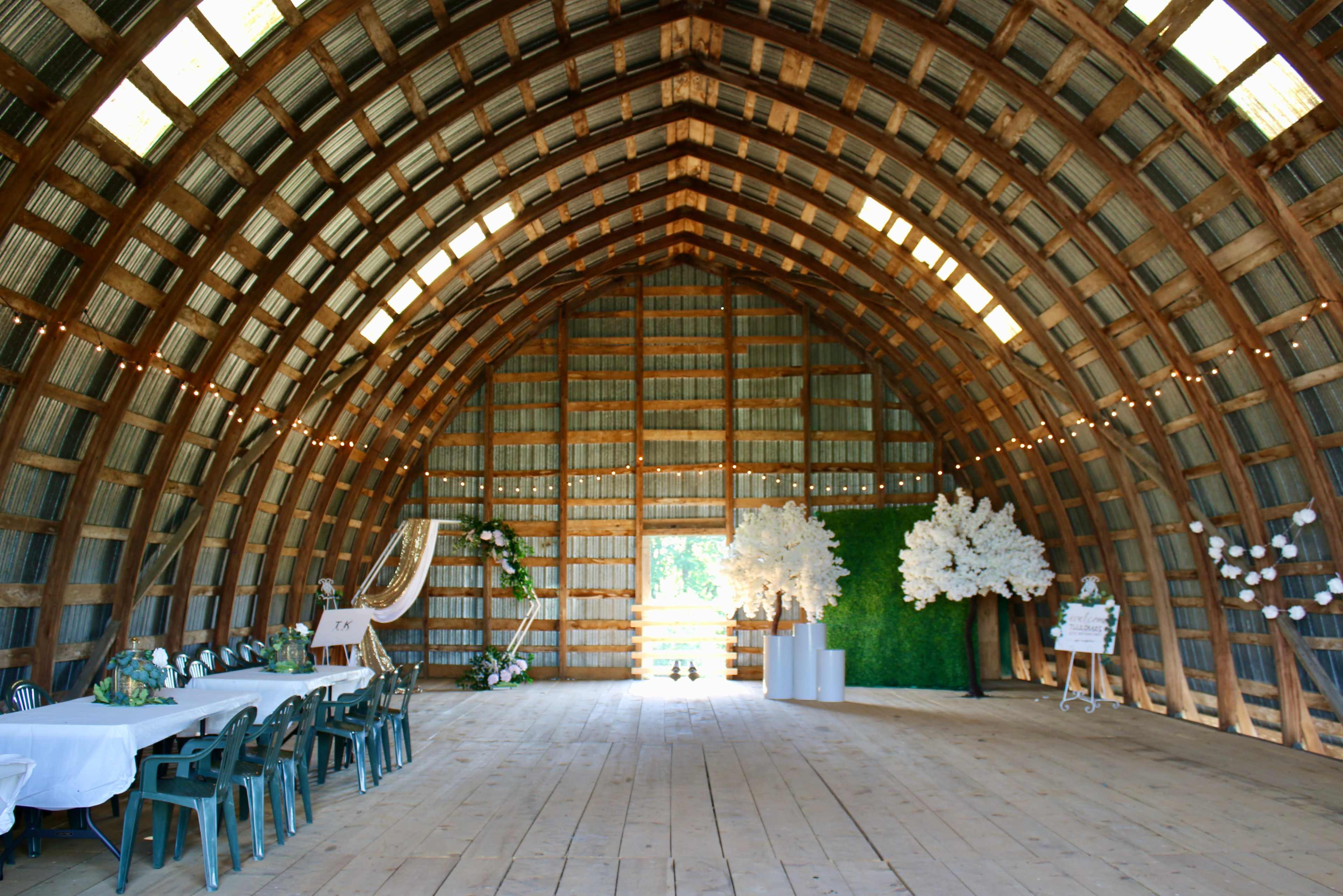 The interior of a barn features wooden beams, a wooden floor, a long table set for dining, and decorative displays of white flowers.