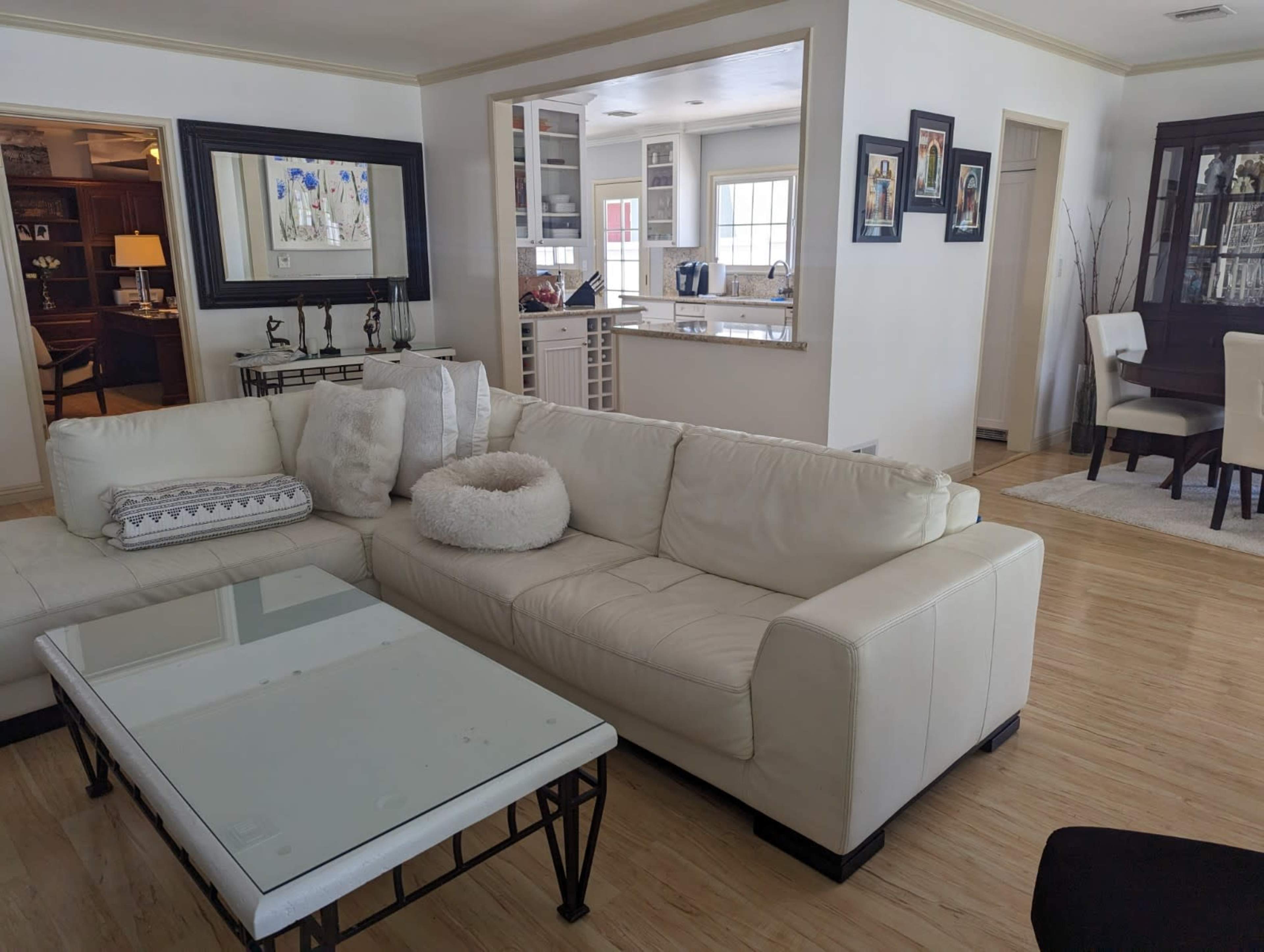 A light-colored living room with a sectional sofa, a glass-top coffee table, and an open layout leading to a dining area.
