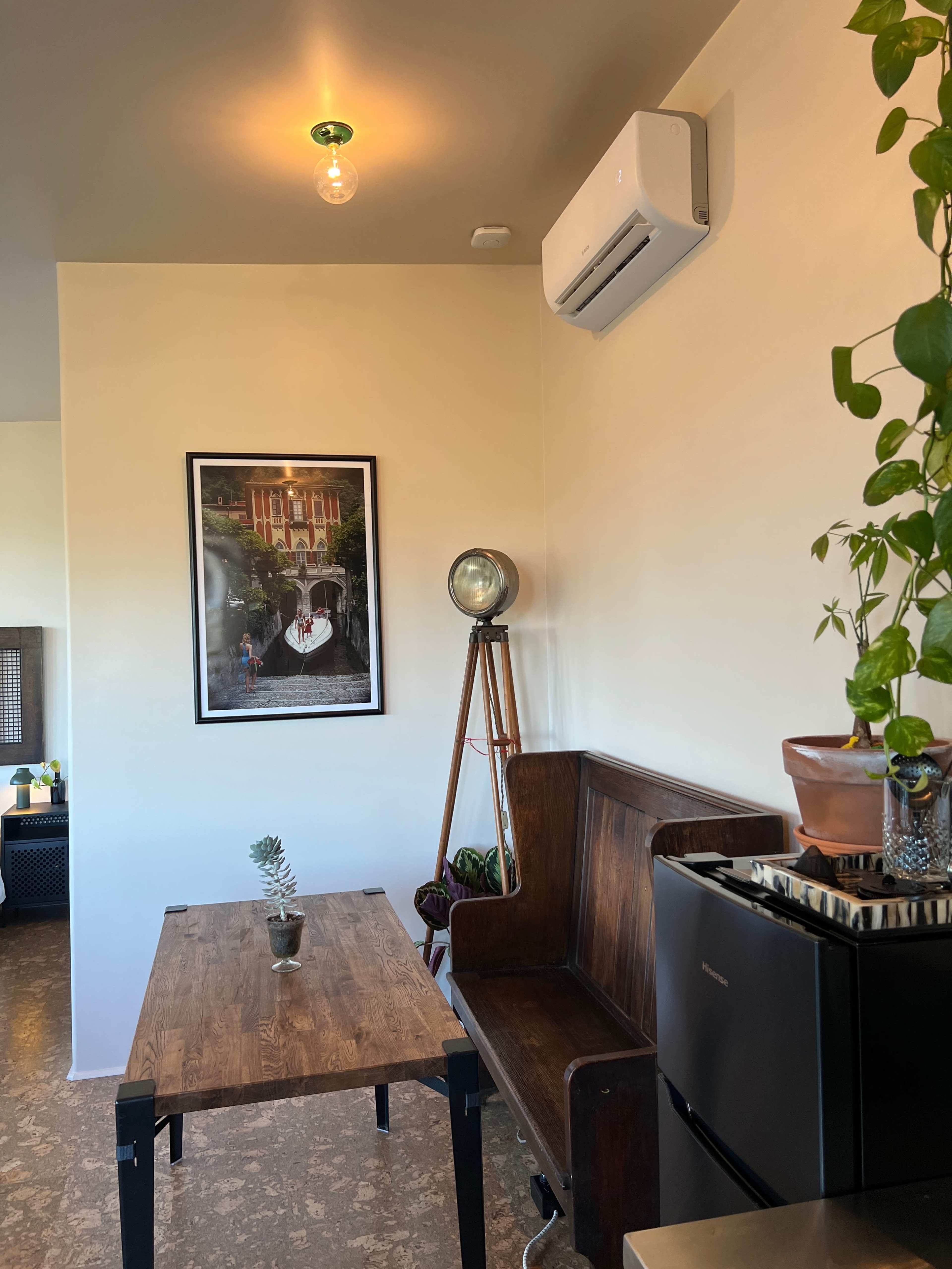 The image shows a cozy corner of a room featuring a wooden bench, a small table, an air conditioning unit, and a framed picture on the wall.