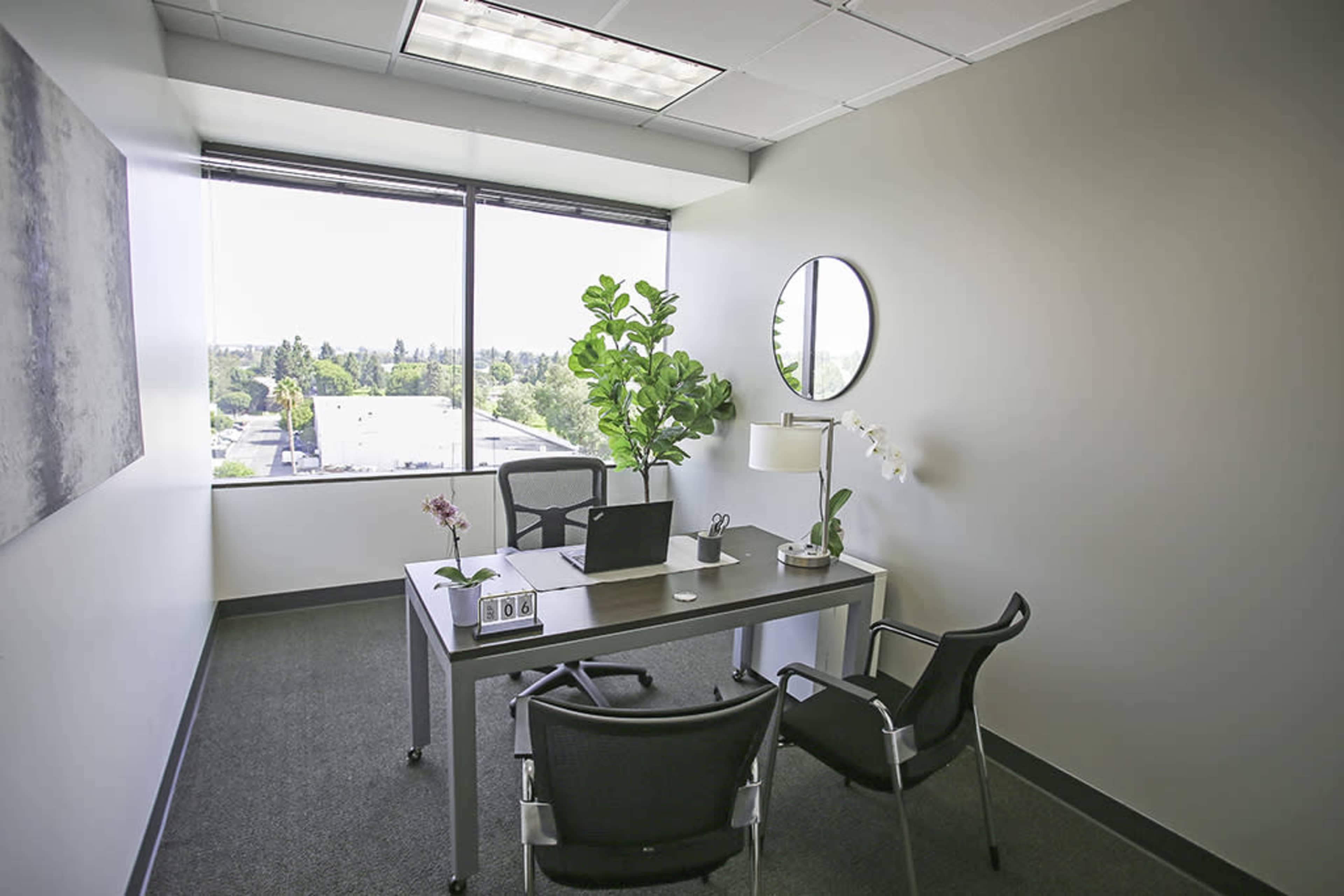 A small office features a desk with a laptop, a lamp, and a plant, accompanied by two chairs and a large window overlooking a landscape.