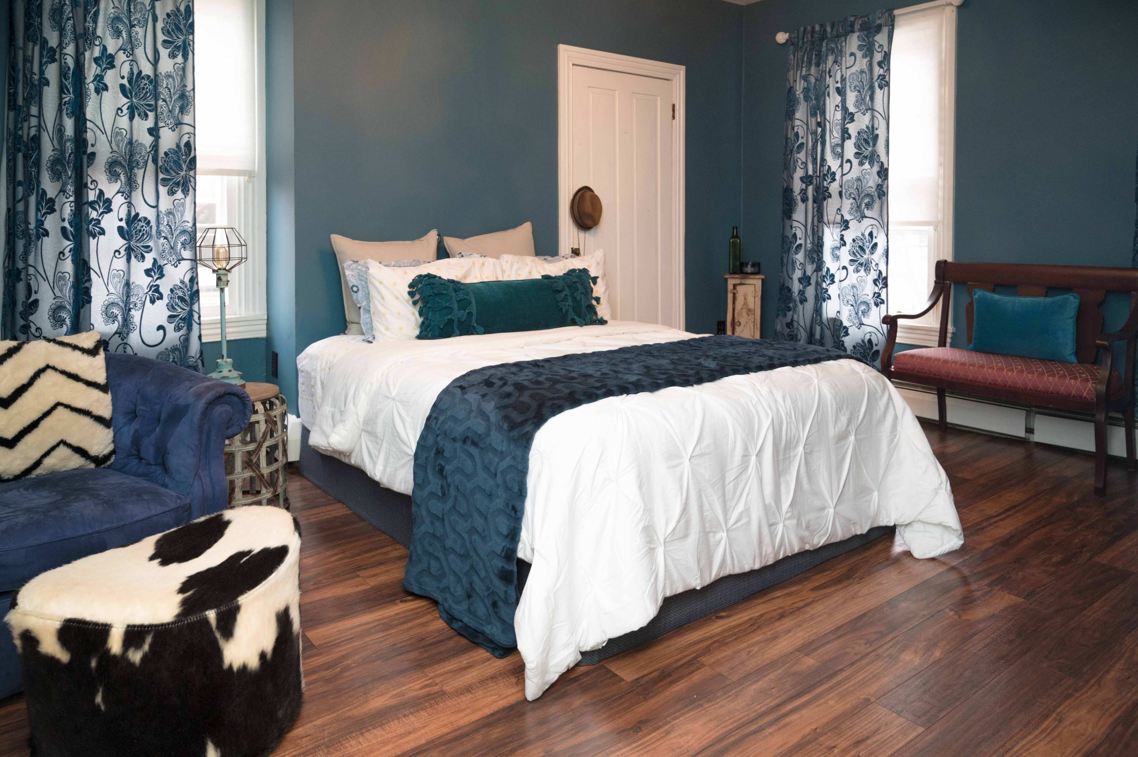 The image shows a cozy bedroom with a bed covered in white and blue bedding, a dark blue accent wall, and various decorative furnishings including a patterned chair and a cowhide ottoman.