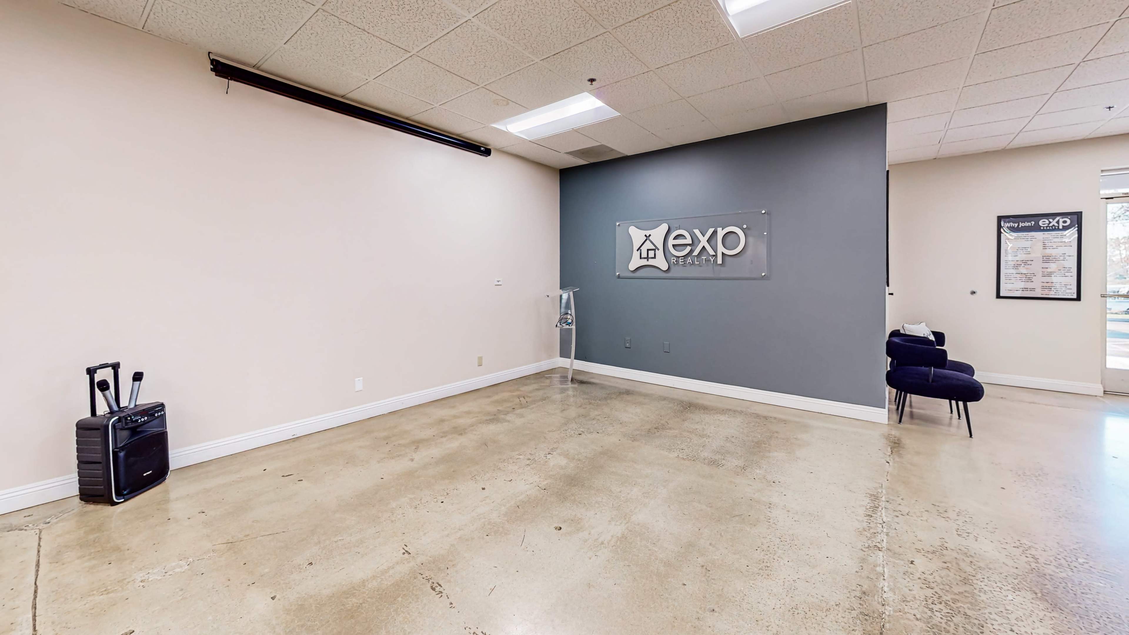 The image shows an empty office space with a polished concrete floor, a wall displaying the logo of exp Realty, and a small seating area with a chair.