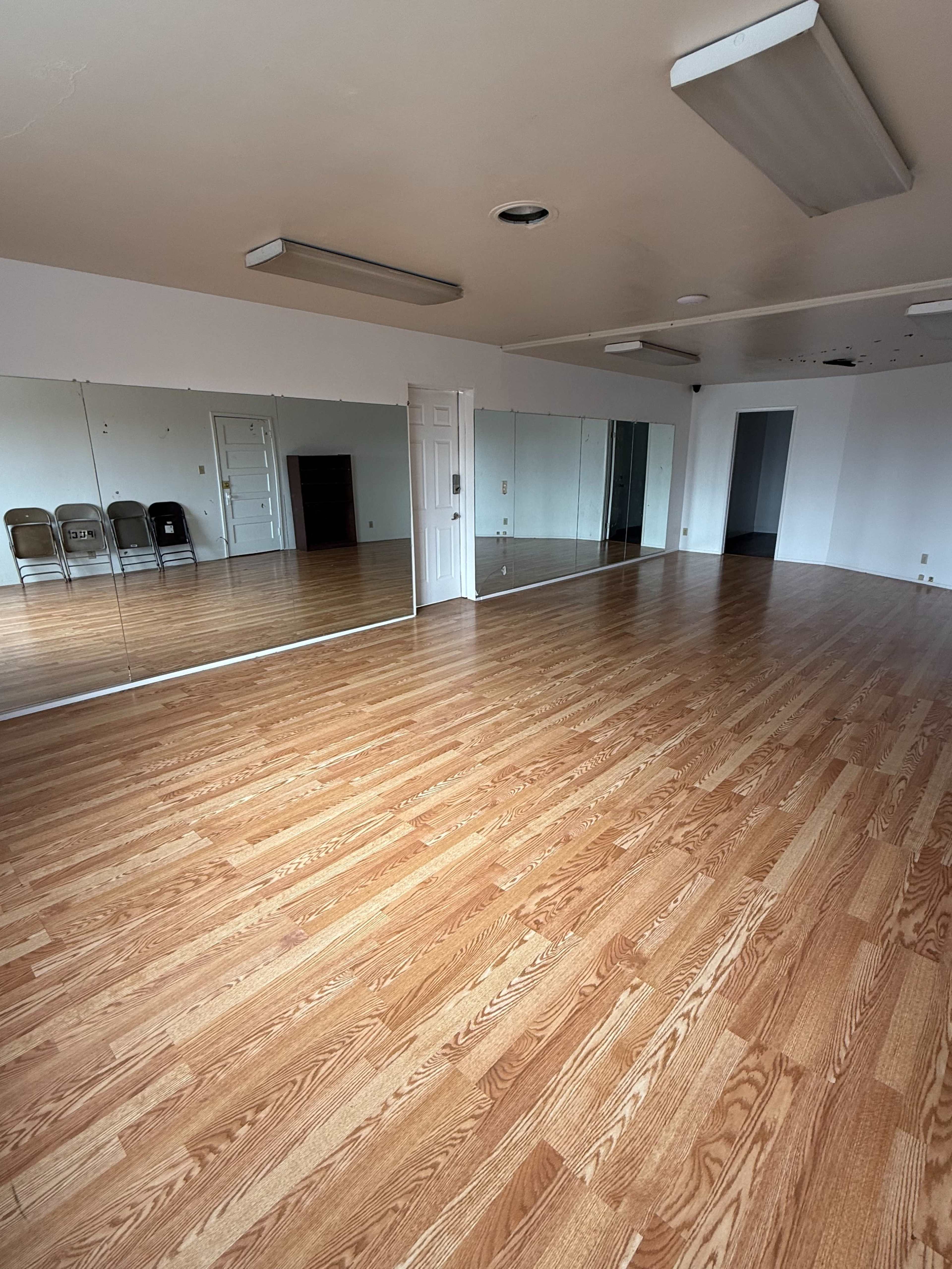 The image shows an empty room with wooden flooring, large mirrors on one wall, and a few folding chairs in the background.