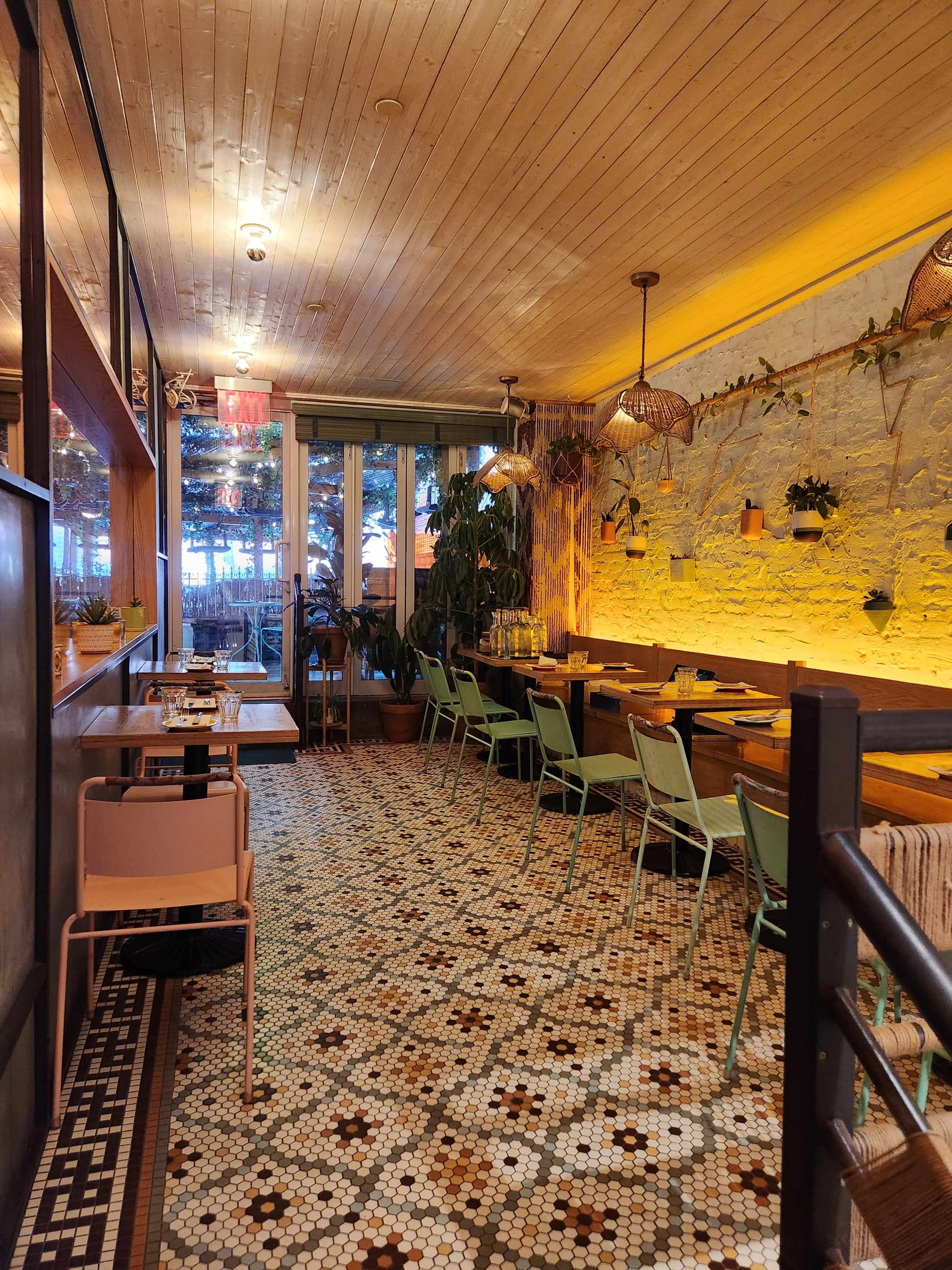 A cozy restaurant interior featuring a tiled floor, wooden ceiling, and green chairs arranged along wooden tables, with plants adorning the walls and hanging light fixtures.