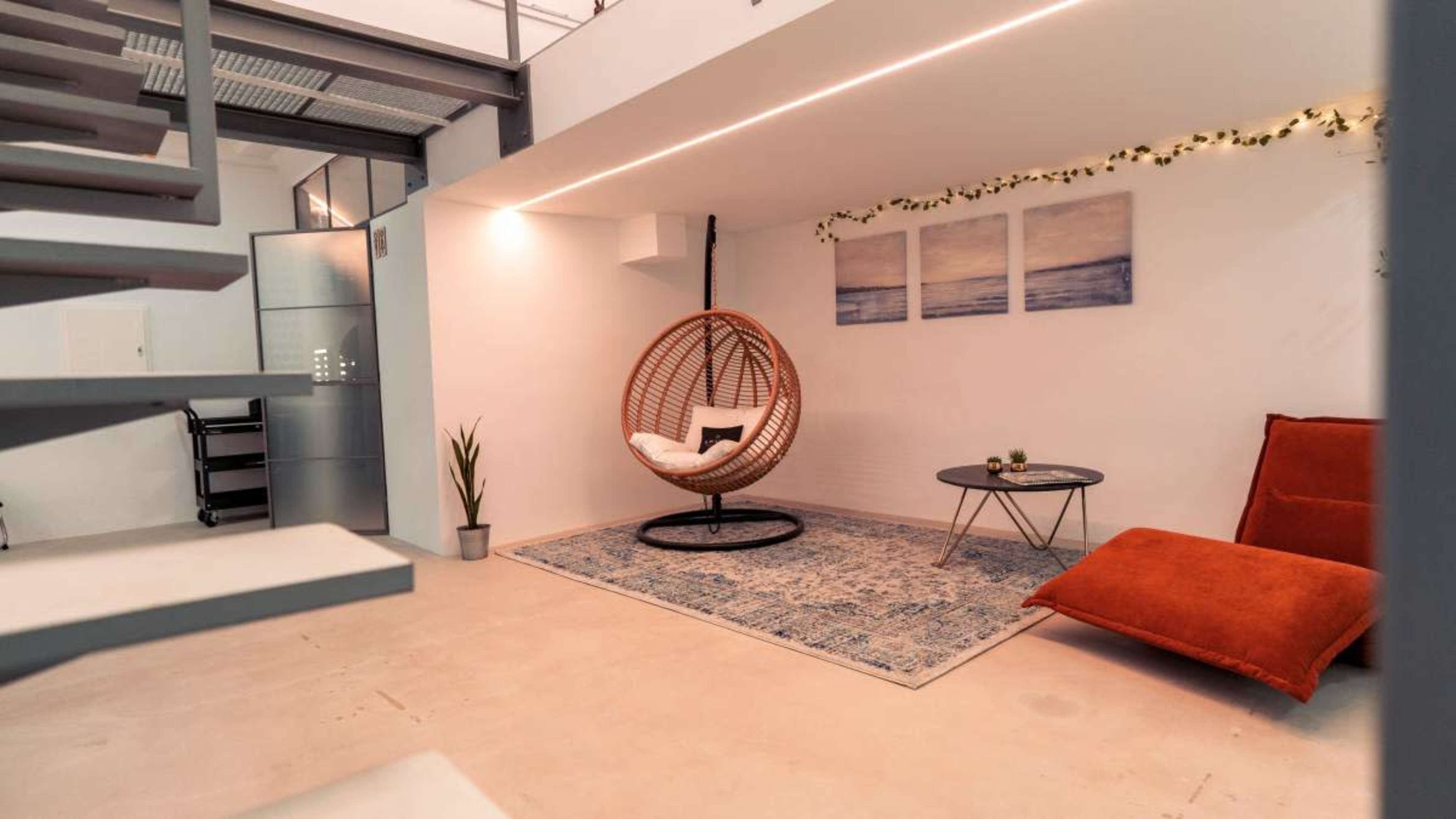 The image shows a modern, minimalist indoor space featuring a hanging chair, a small round table, and a red lounge chair on a decorative rug.