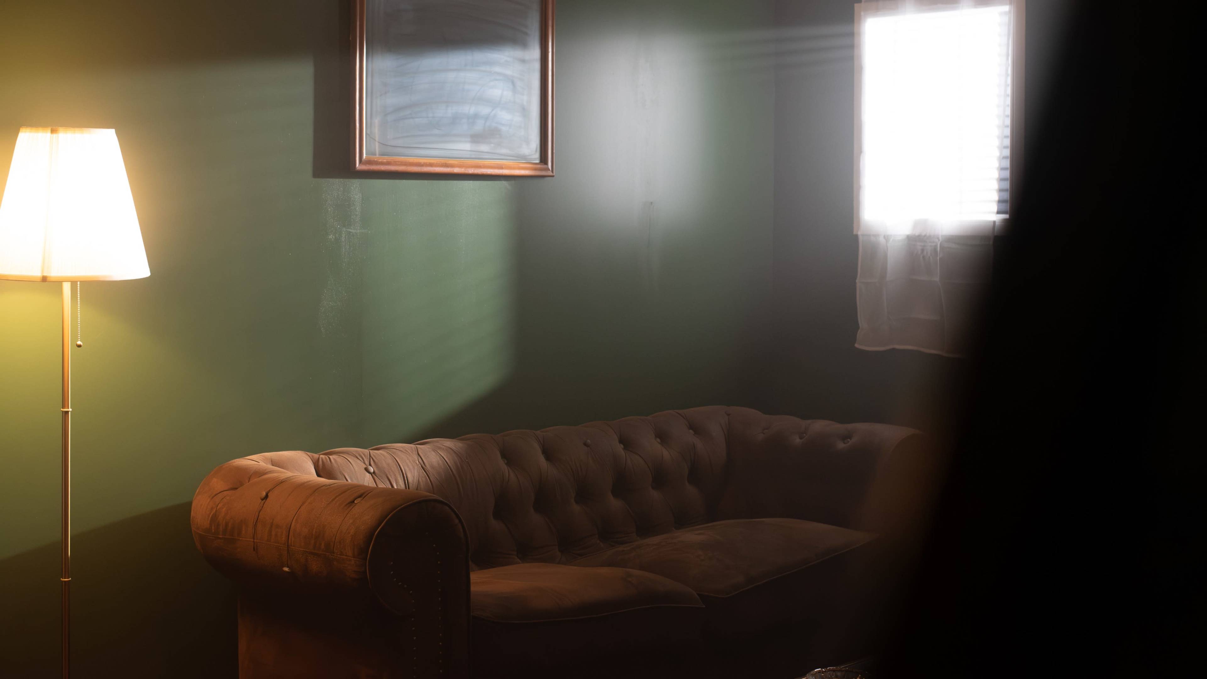 A brown tufted sofa sits in a dimly lit room with a green wall, a floor lamp, and a framed painting above it.