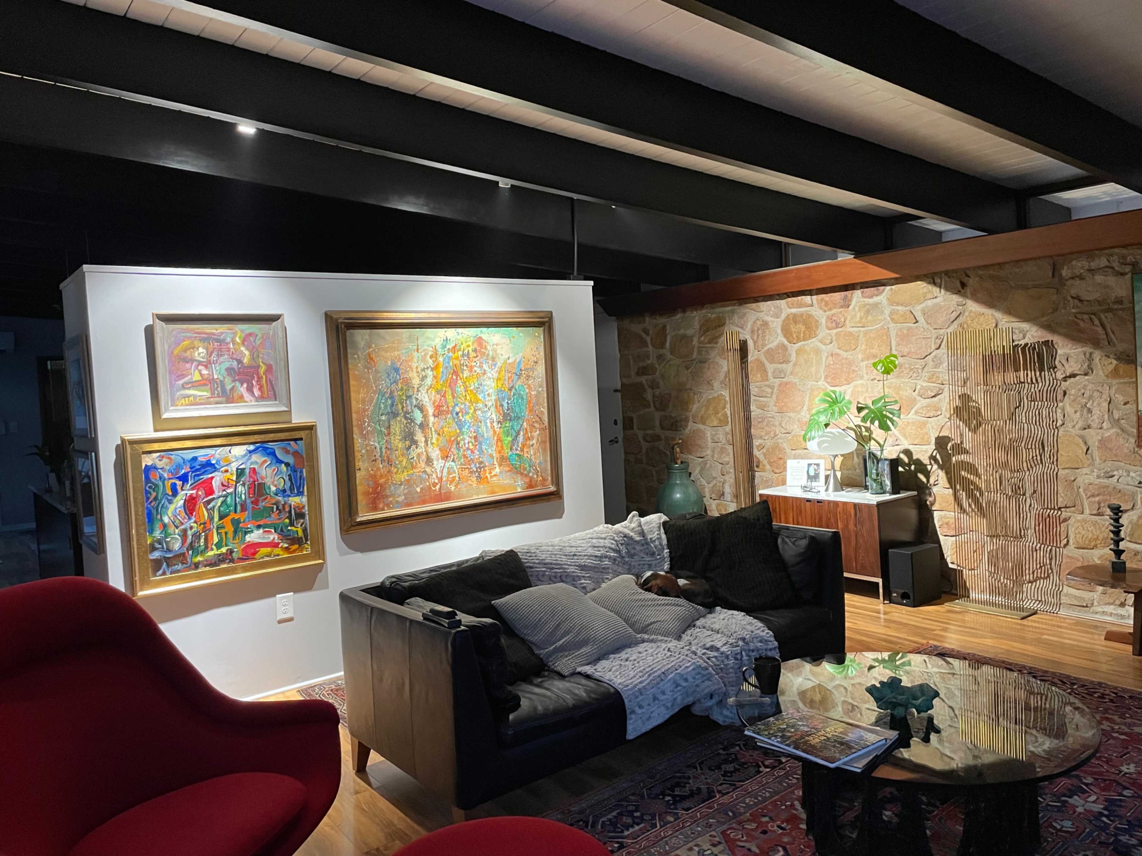 A living room features a black couch with a throw blanket, a round glass coffee table, colorful paintings on the wall, and a stone accent wall.