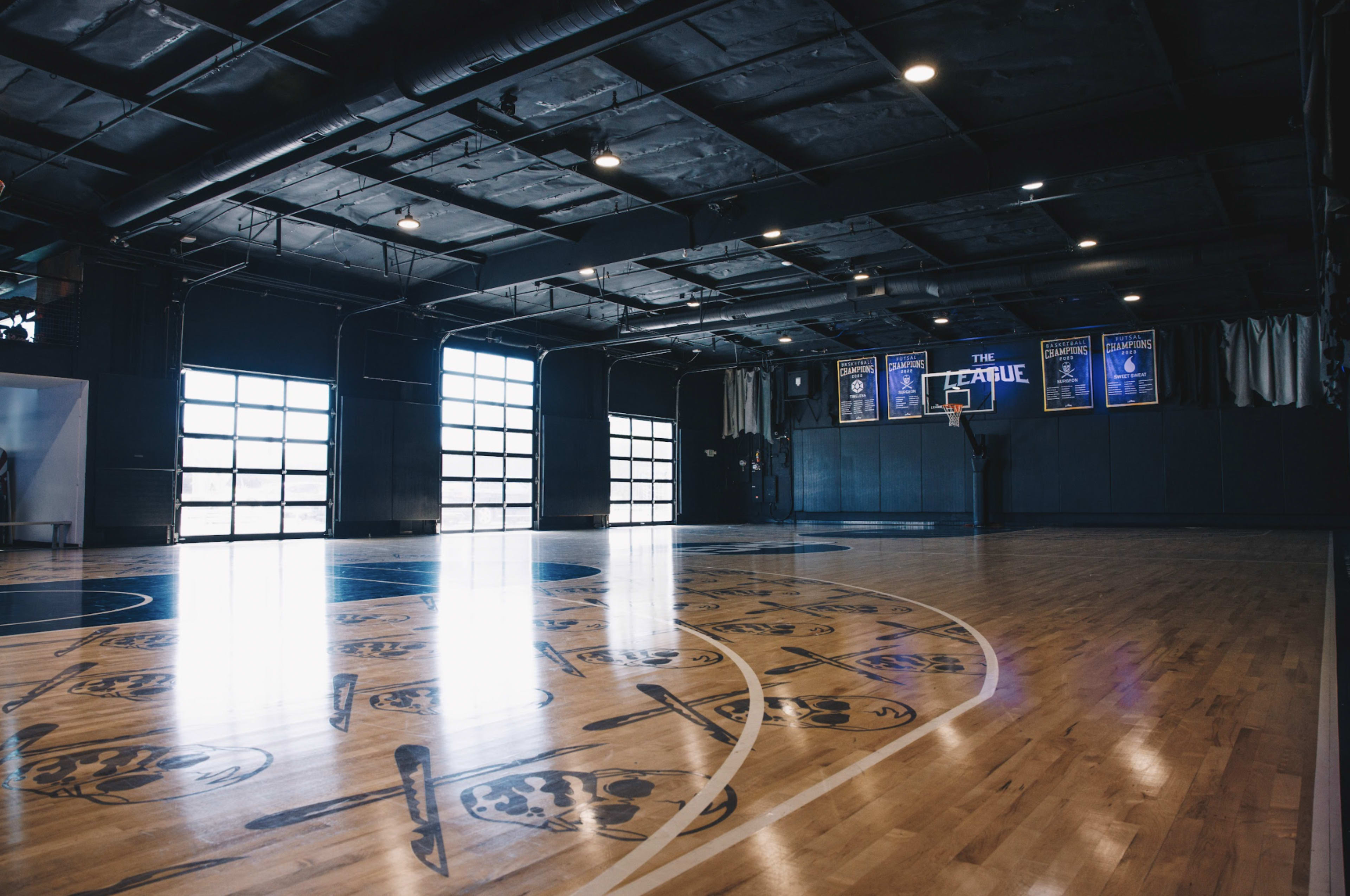 The image shows an indoor basketball court with large windows, polished wooden floors, and a basketball hoop at one end.