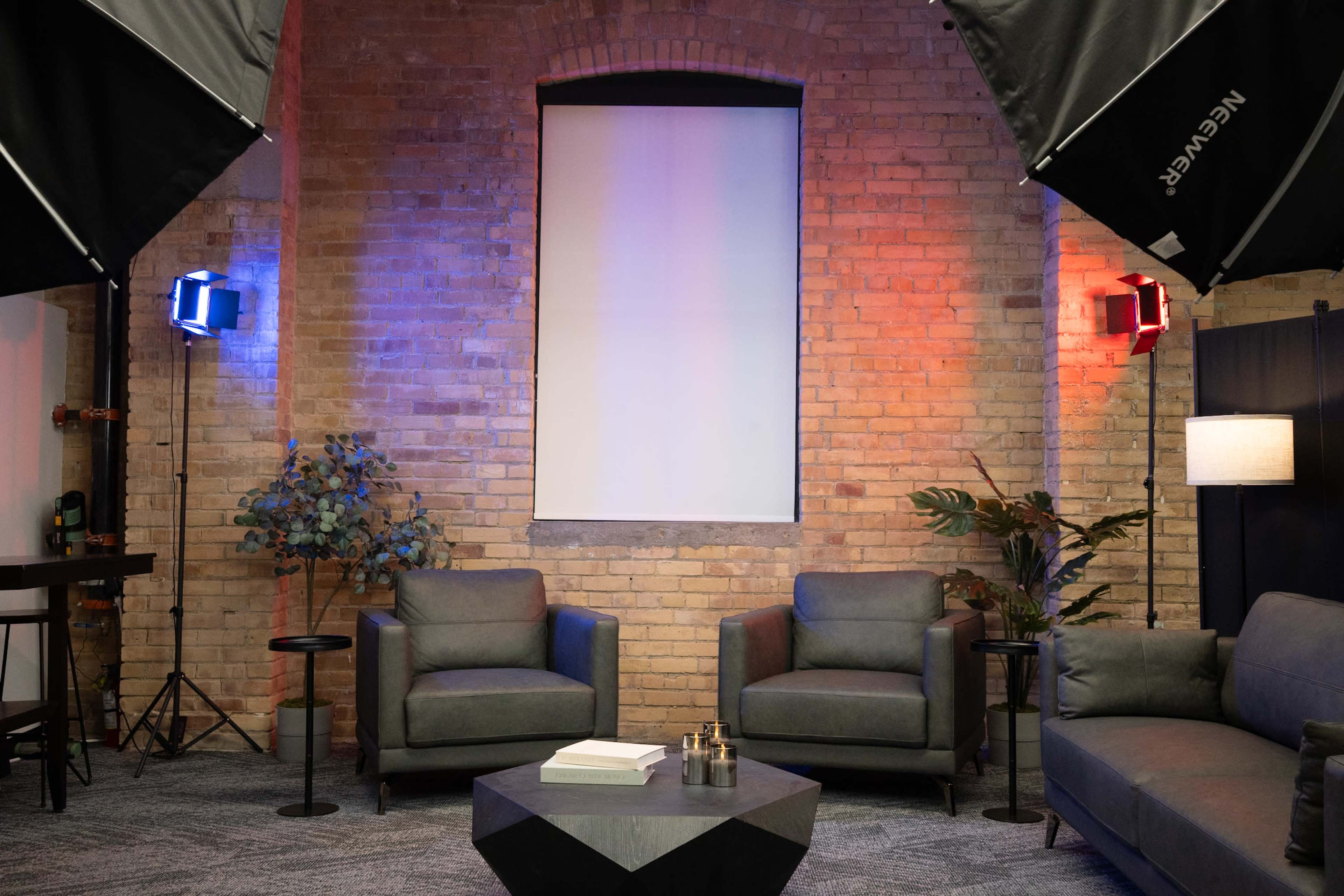 The image shows a modern lounge area with two gray sofas, a coffee table, and a backdrop illuminated by colored lights against a brick wall.