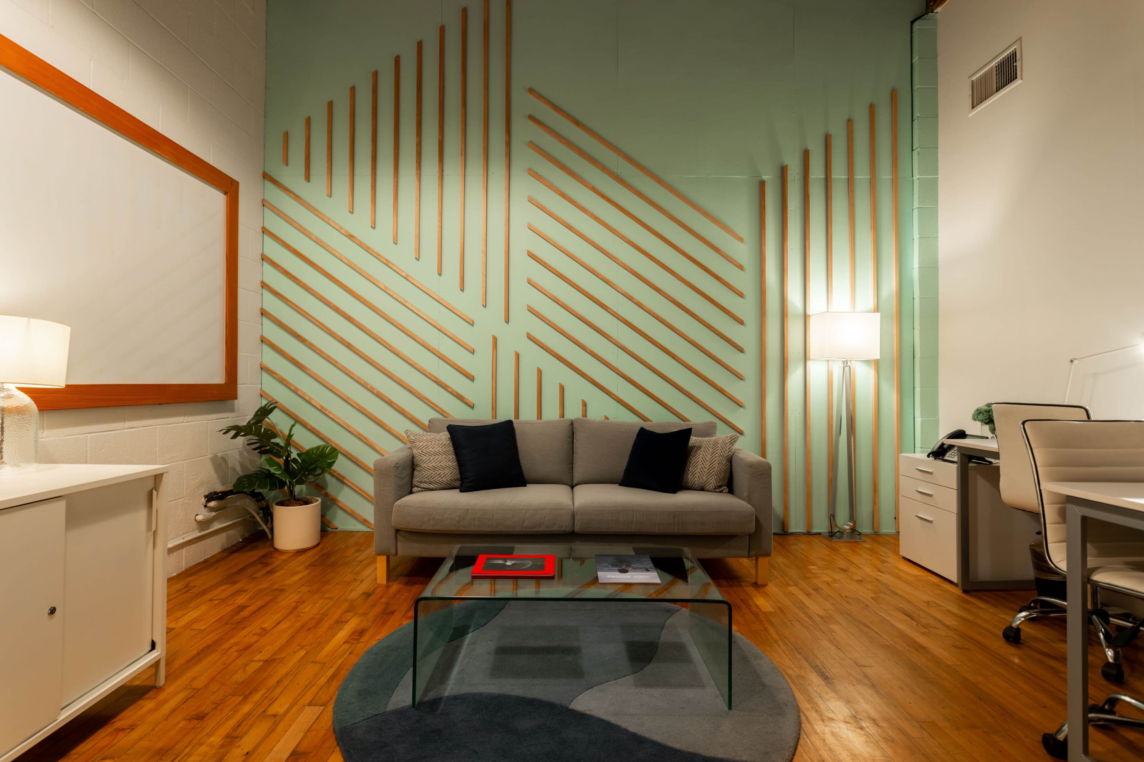 The image shows a modern lounge area featuring a gray sofa with black pillows, a glass coffee table, and a work desk, all set against a mint green wall with diagonal wooden slats.