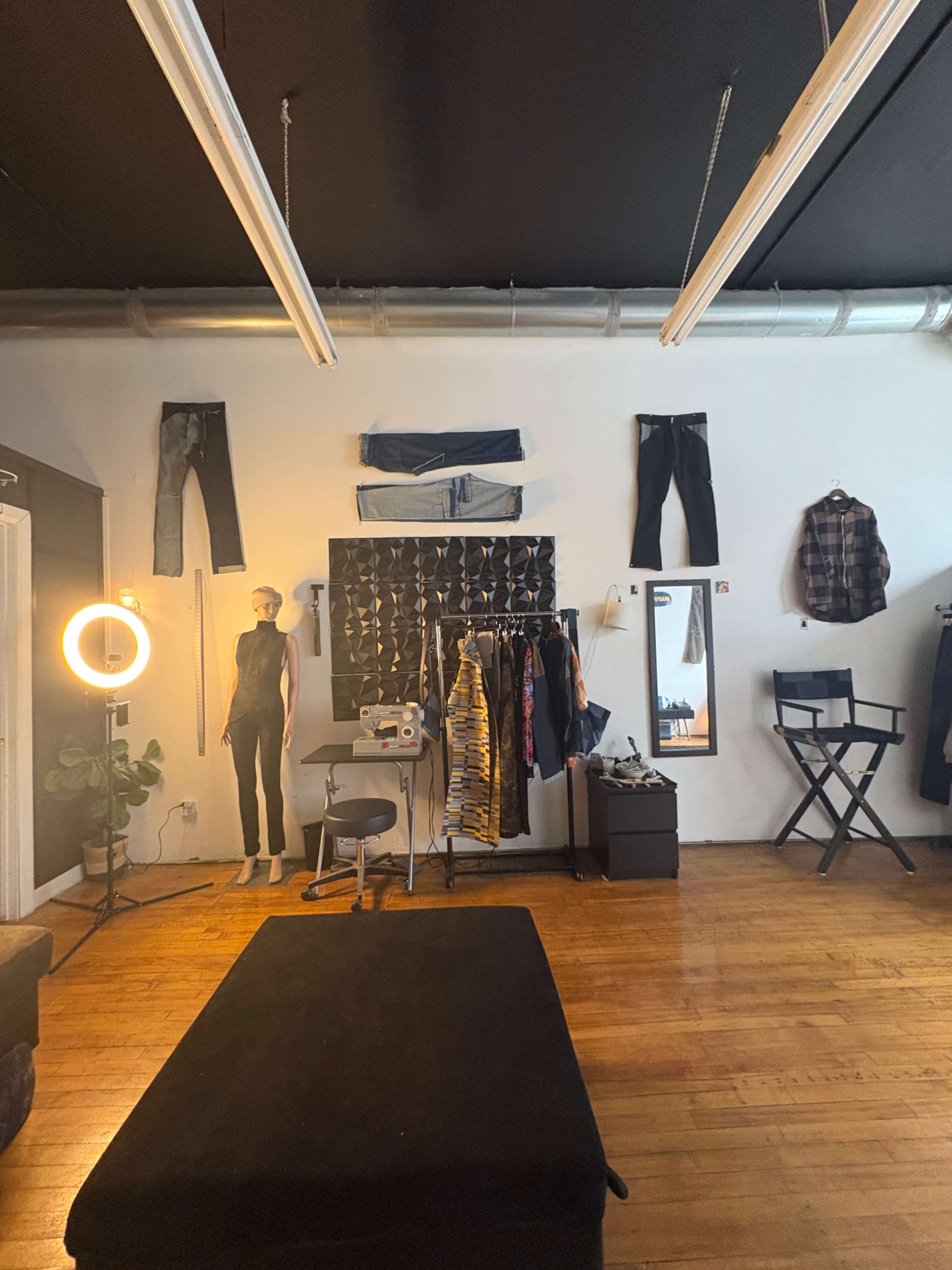 The image depicts a clothing studio featuring a mannequin dressed in a jumpsuit, various pairs of jeans hanging on the wall, a sewing machine, and a director's chair in a well-lit space with wooden flooring.
