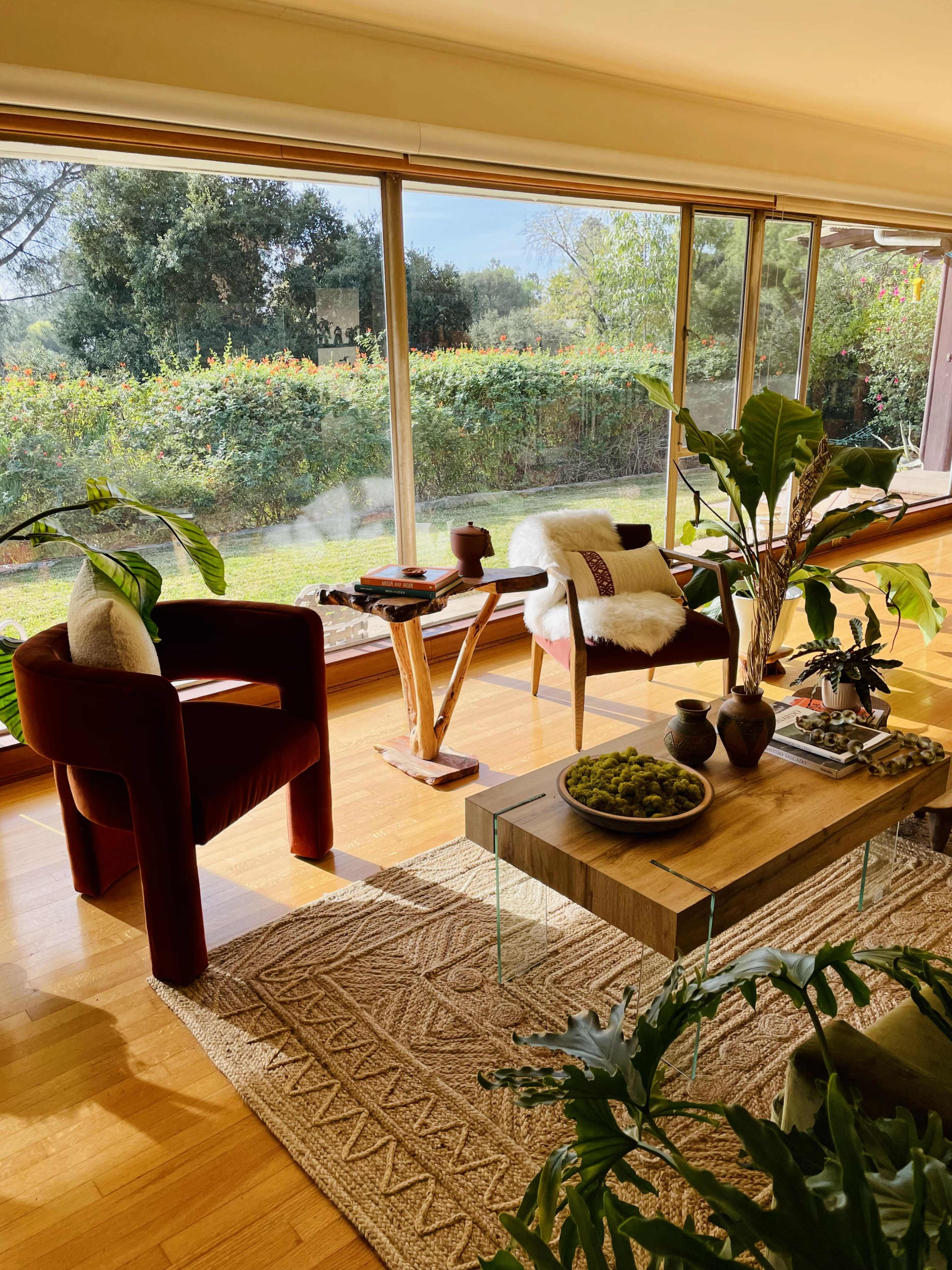 The image shows a well-lit living room with two chairs, a coffee table, and large windows overlooking a lush green garden.