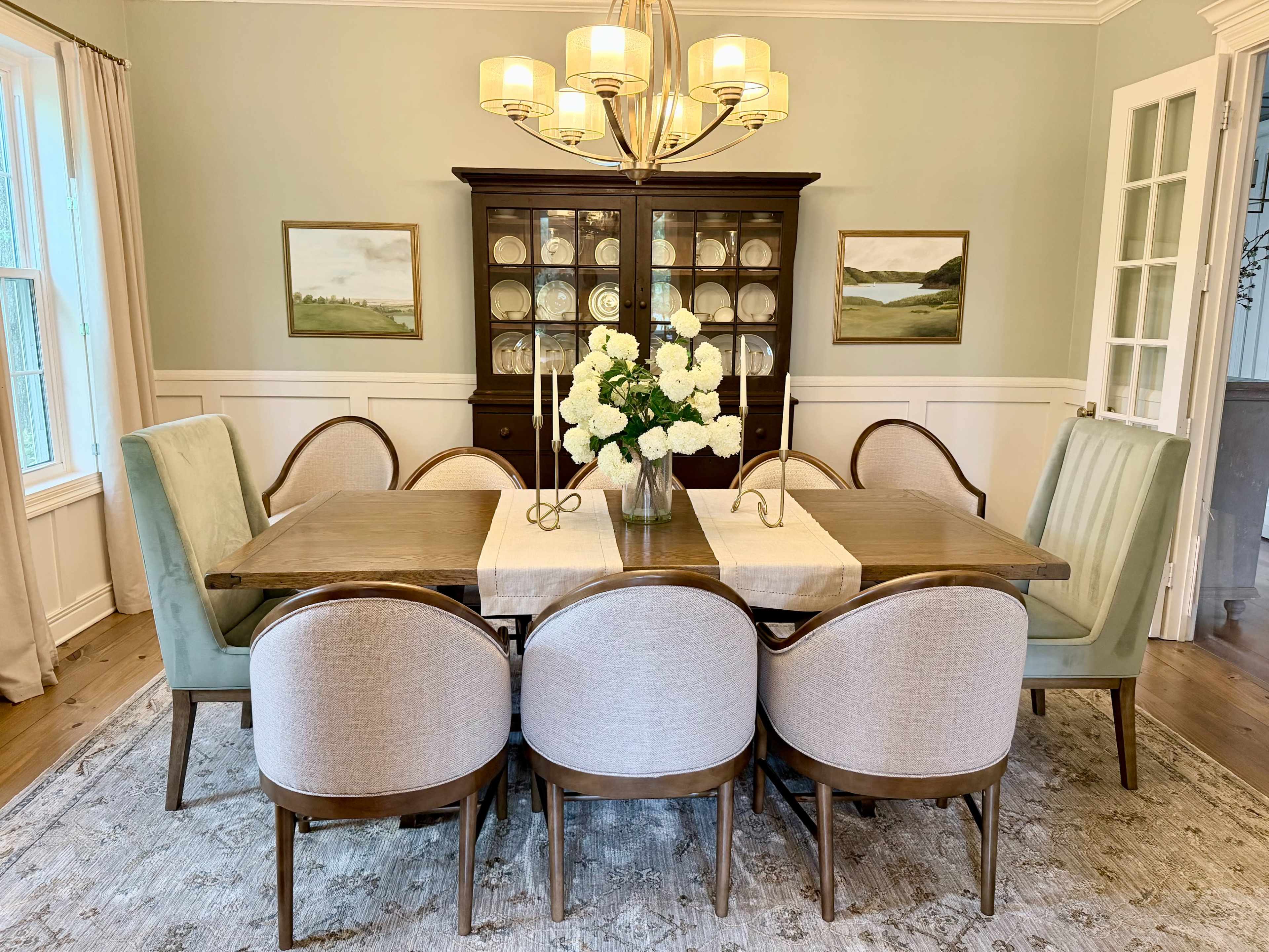 A brightly lit dining room features a large wooden table surrounded by upholstered chairs, with a vase of white flowers in the center and a china cabinet against the wall.