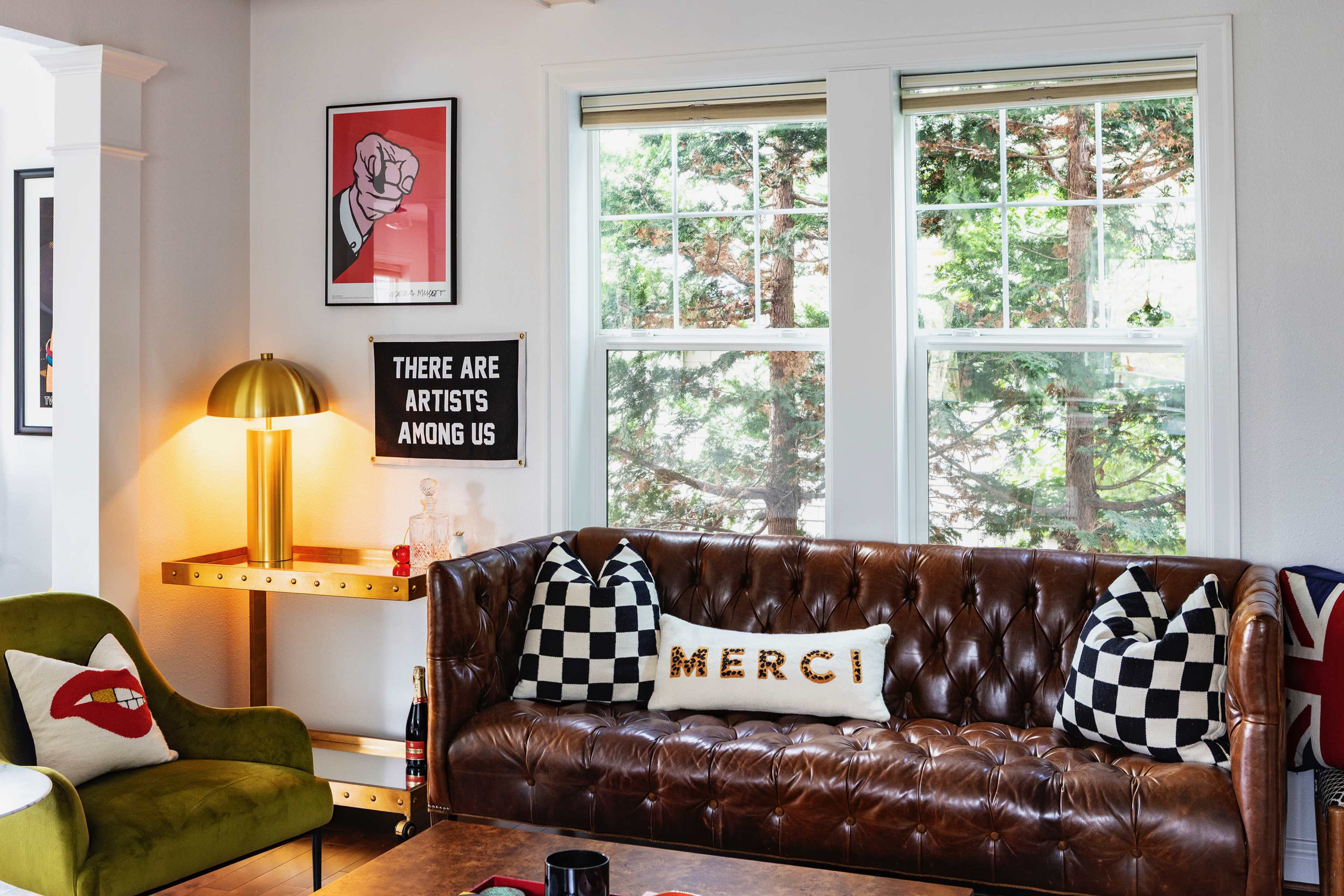 The image shows a cozy living room with a brown leather sofa adorned with patterned pillows, a green armchair, and a decorative lamp beside a window with trees visible outside.