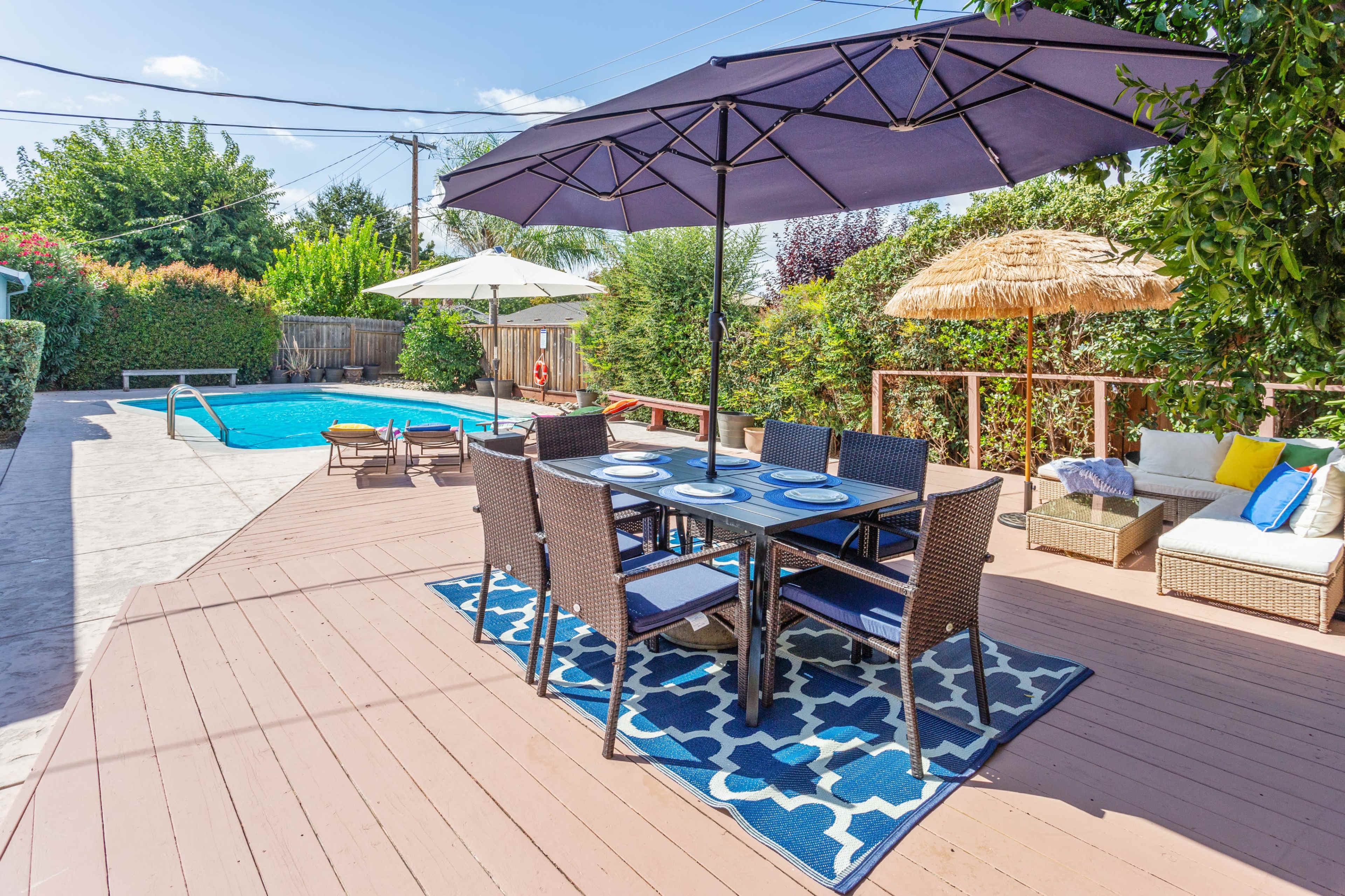 A backyard patio area with a dining table under a large umbrella, surrounded by a swimming pool and various seating arrangements.