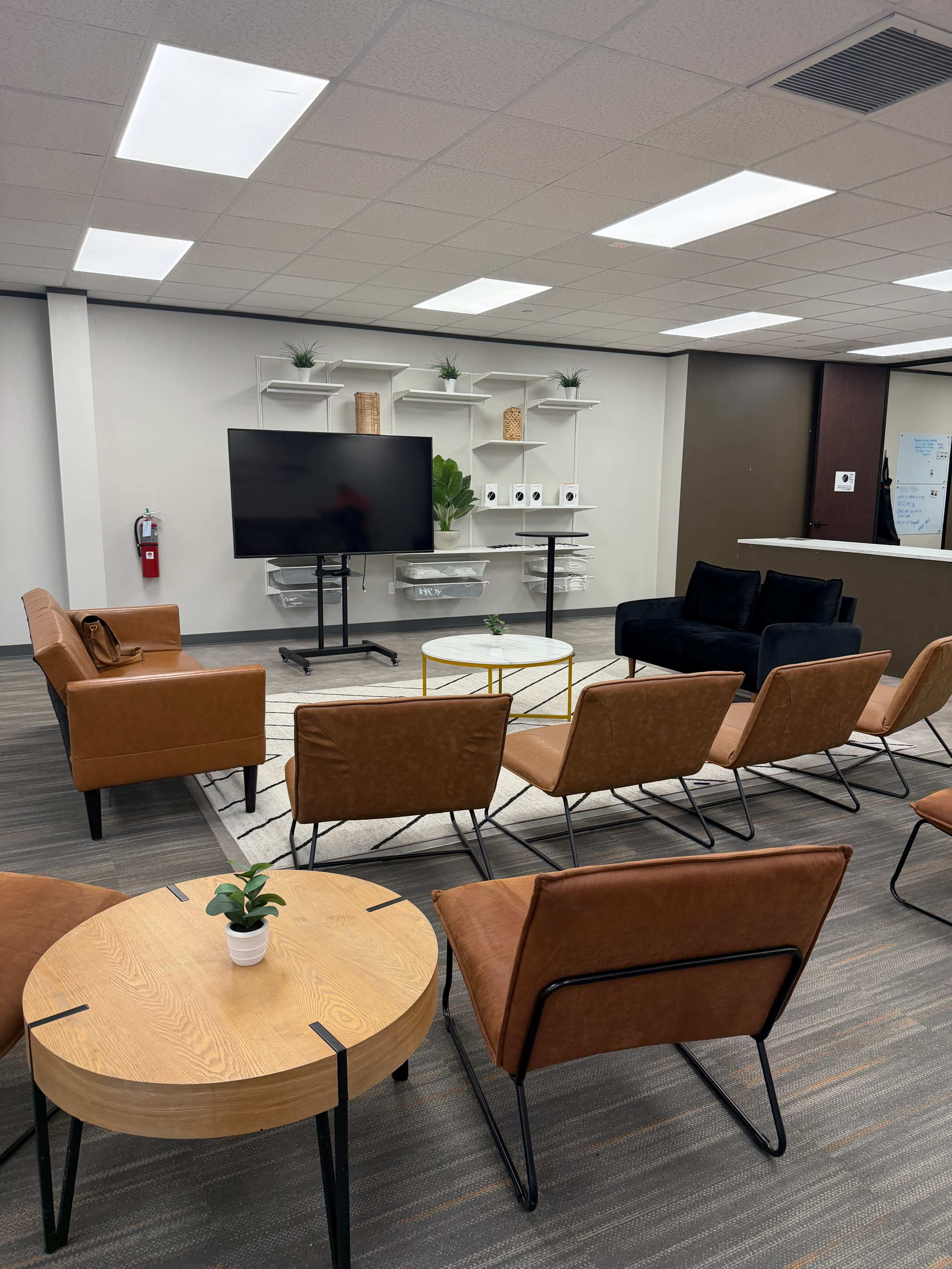 The image shows a modern lounge area with several brown chairs, a coffee table, a television on a stand, and white shelves with plants and decor against a backdrop of gray walls.