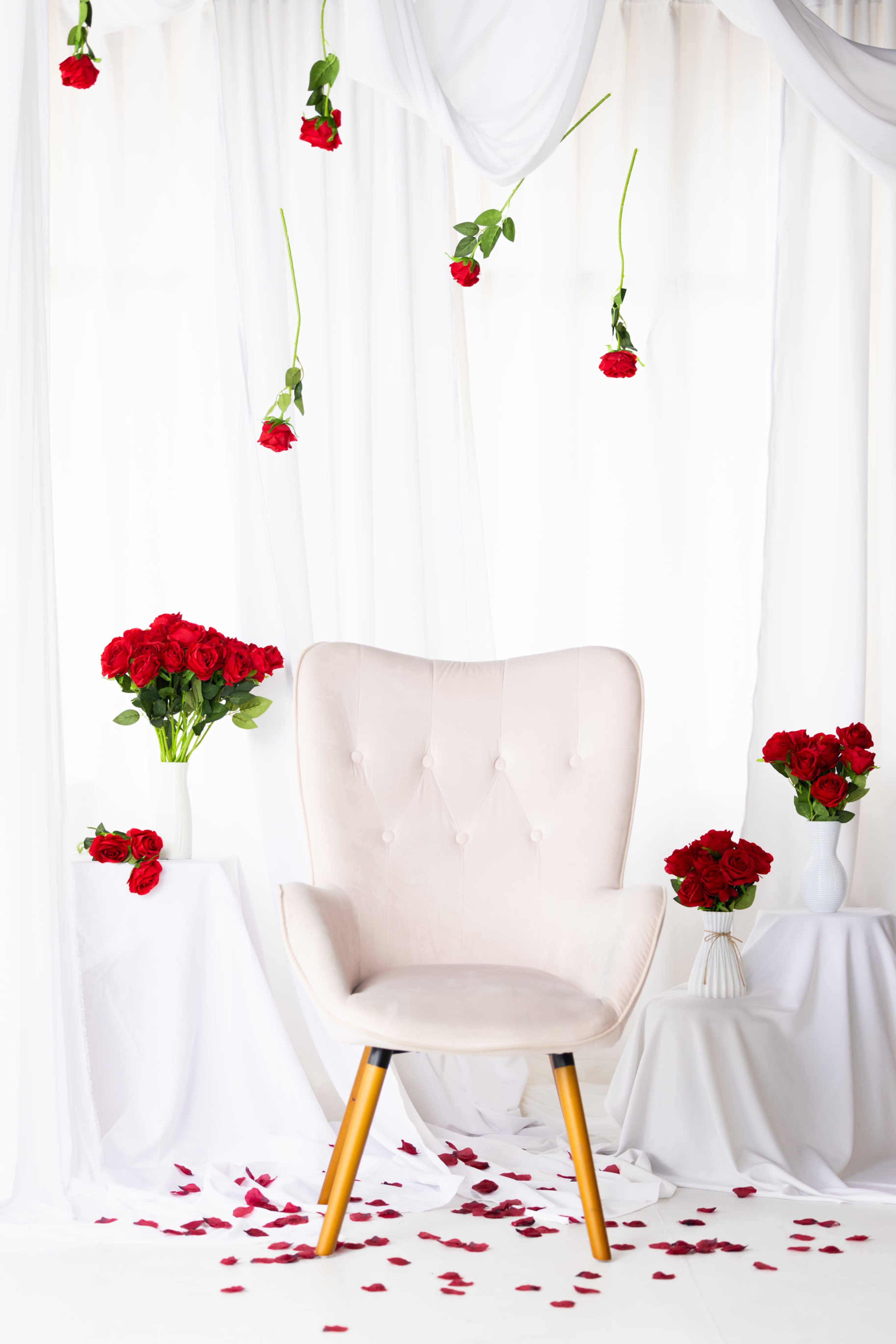 A light-colored chair surrounded by red rose bouquets and scattered rose petals against a backdrop of white drapery.