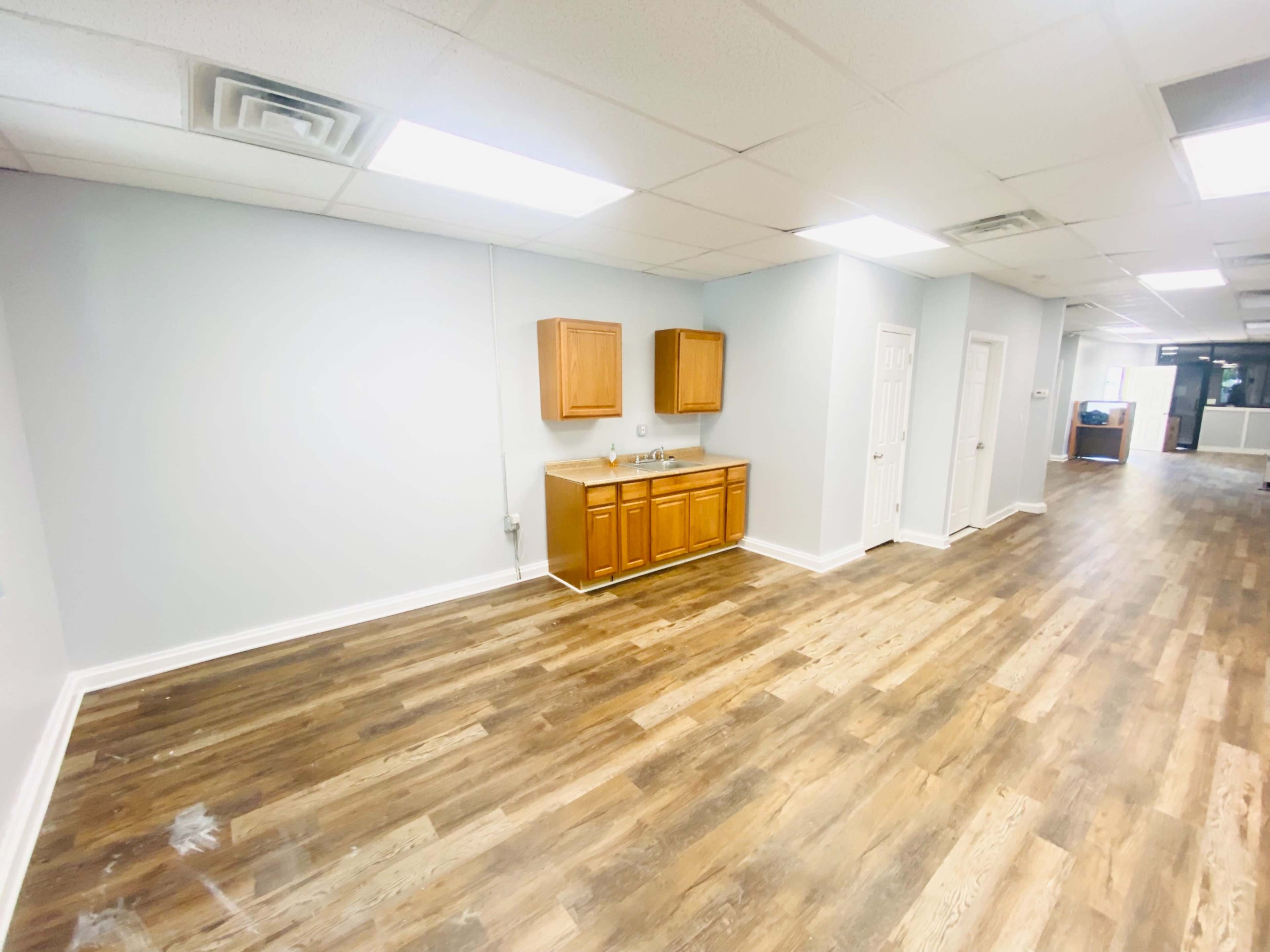 An empty room with wooden flooring, featuring a kitchenette along one wall and doorways leading to other areas.