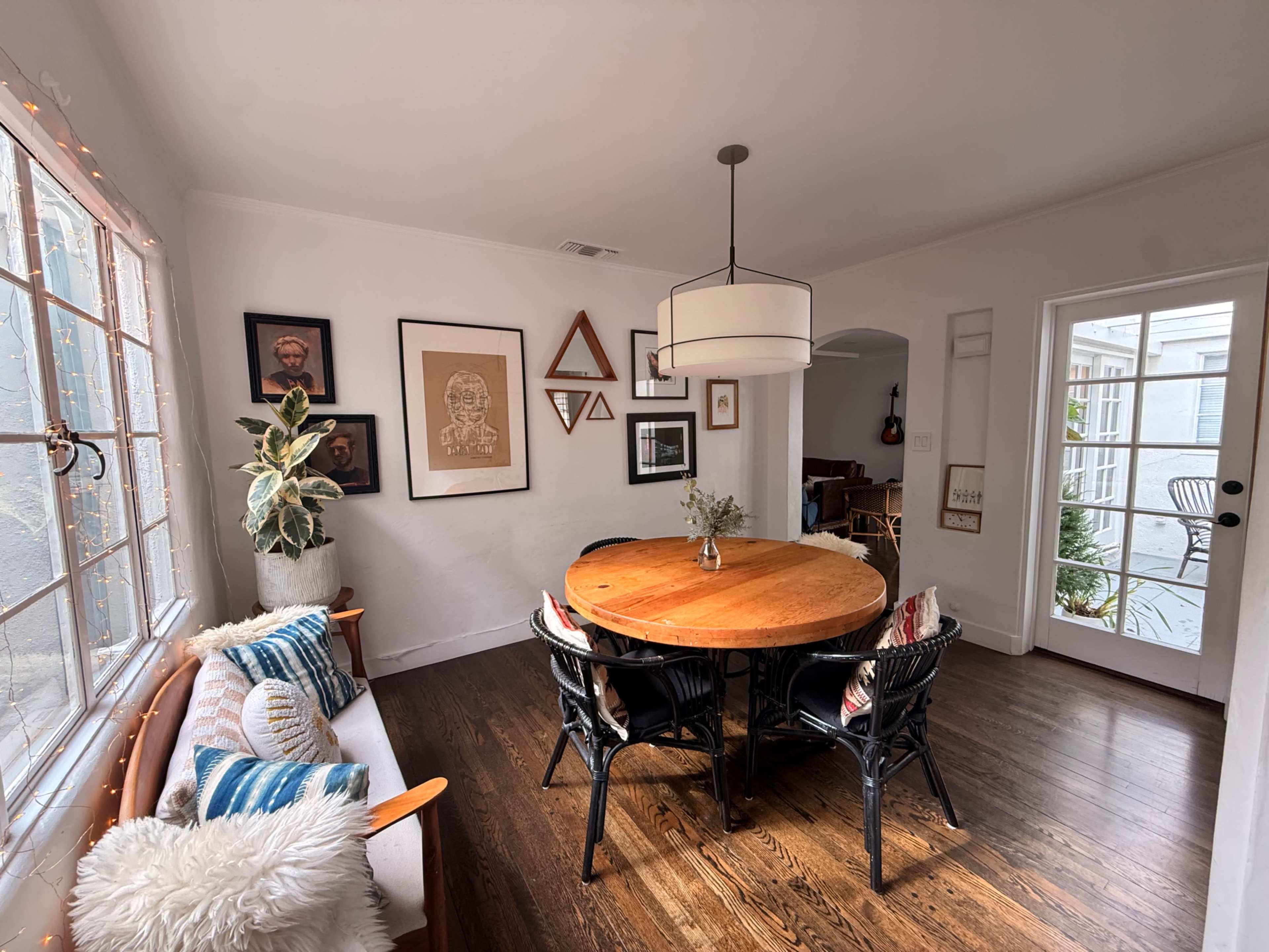 The image depicts a cozy dining area with a round wooden table surrounded by black chairs, accented by framed artworks on the walls and large windows letting in natural light.