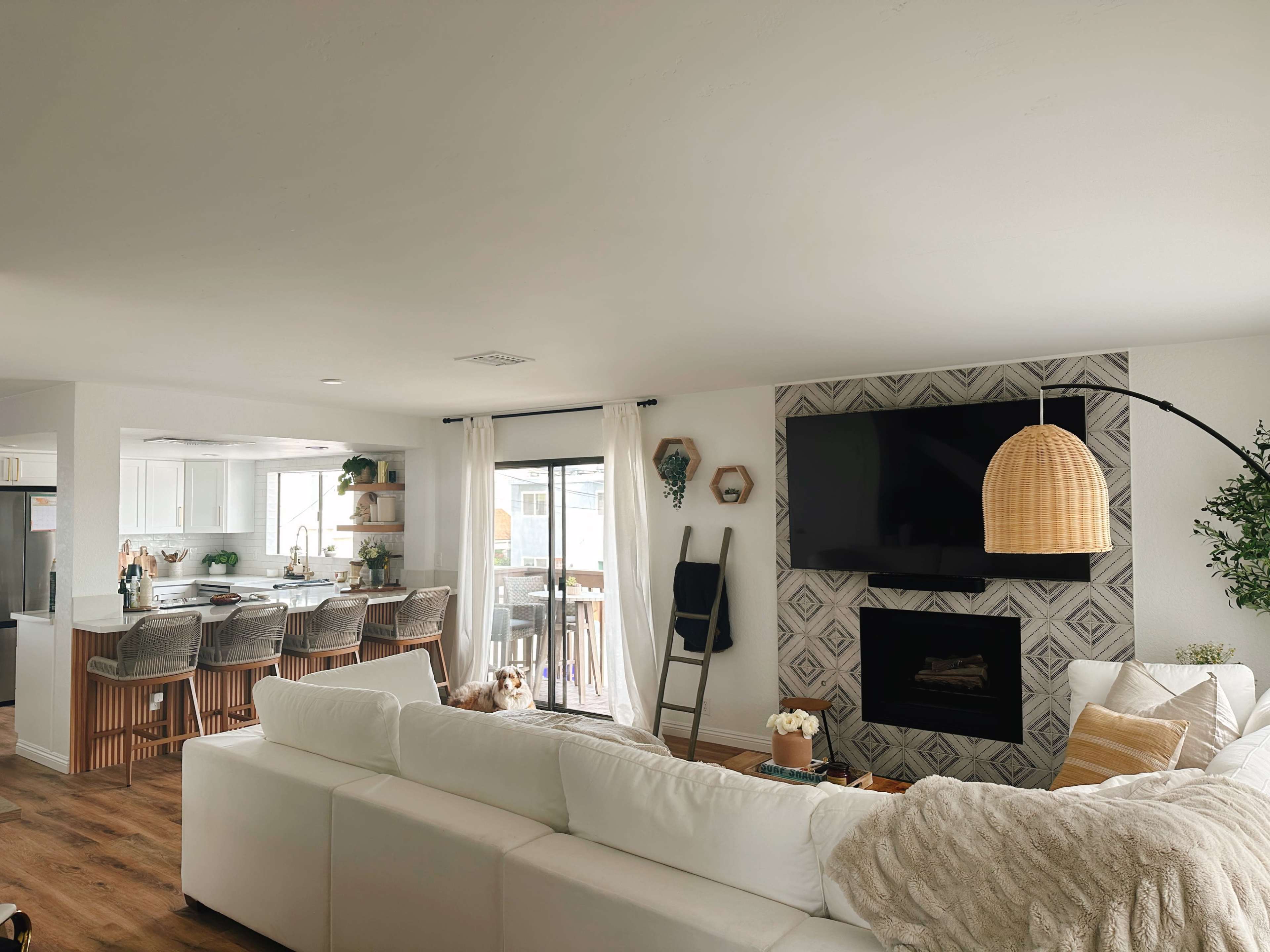 A modern living room with a white sectional sofa facing a wall-mounted TV and a fireplace, while a dining area with bar stools and an open kitchen is visible in the background.