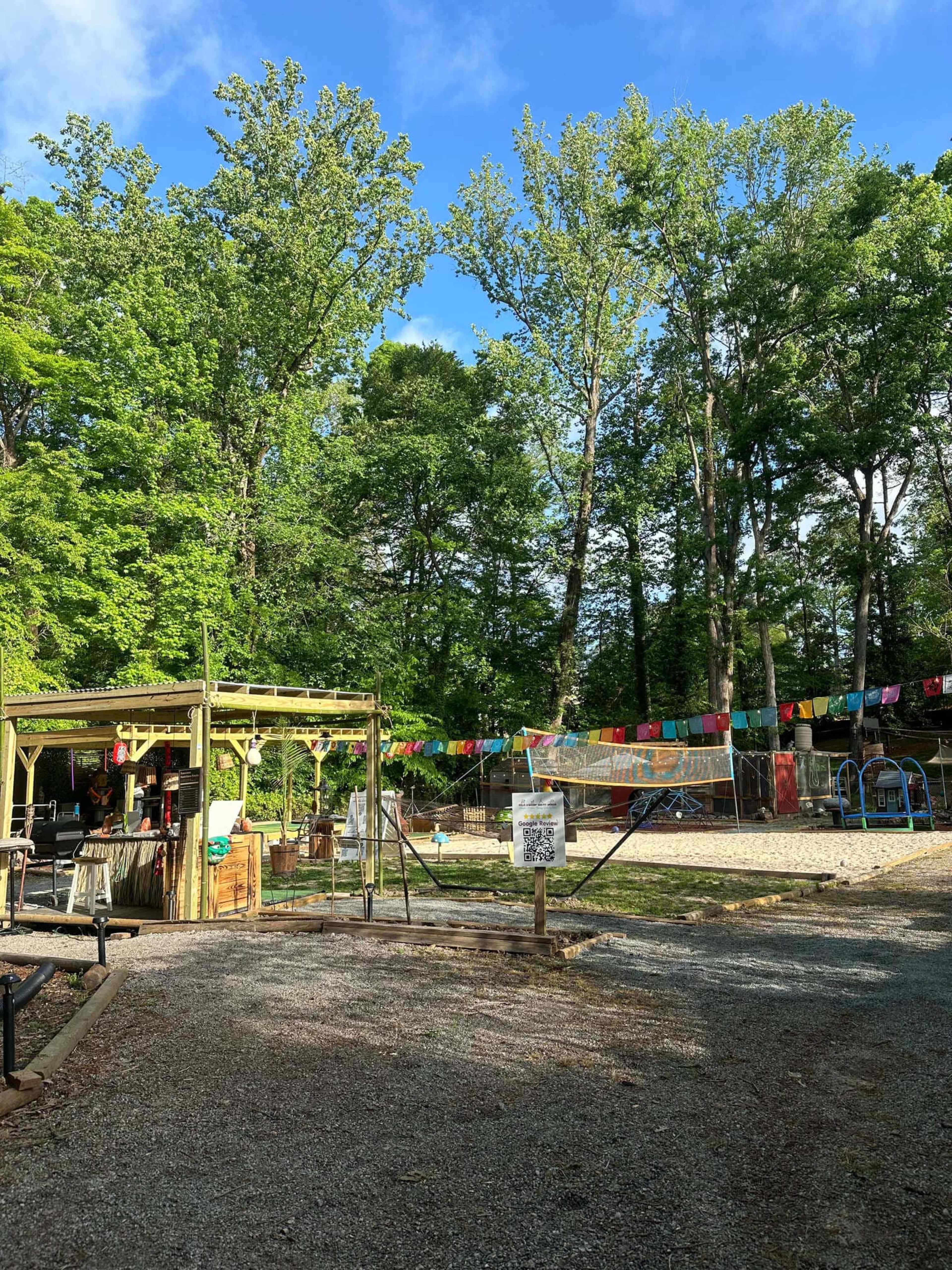 A gravel pathway leads to a gathering area surrounded by tall trees and colorful flags strung between them.