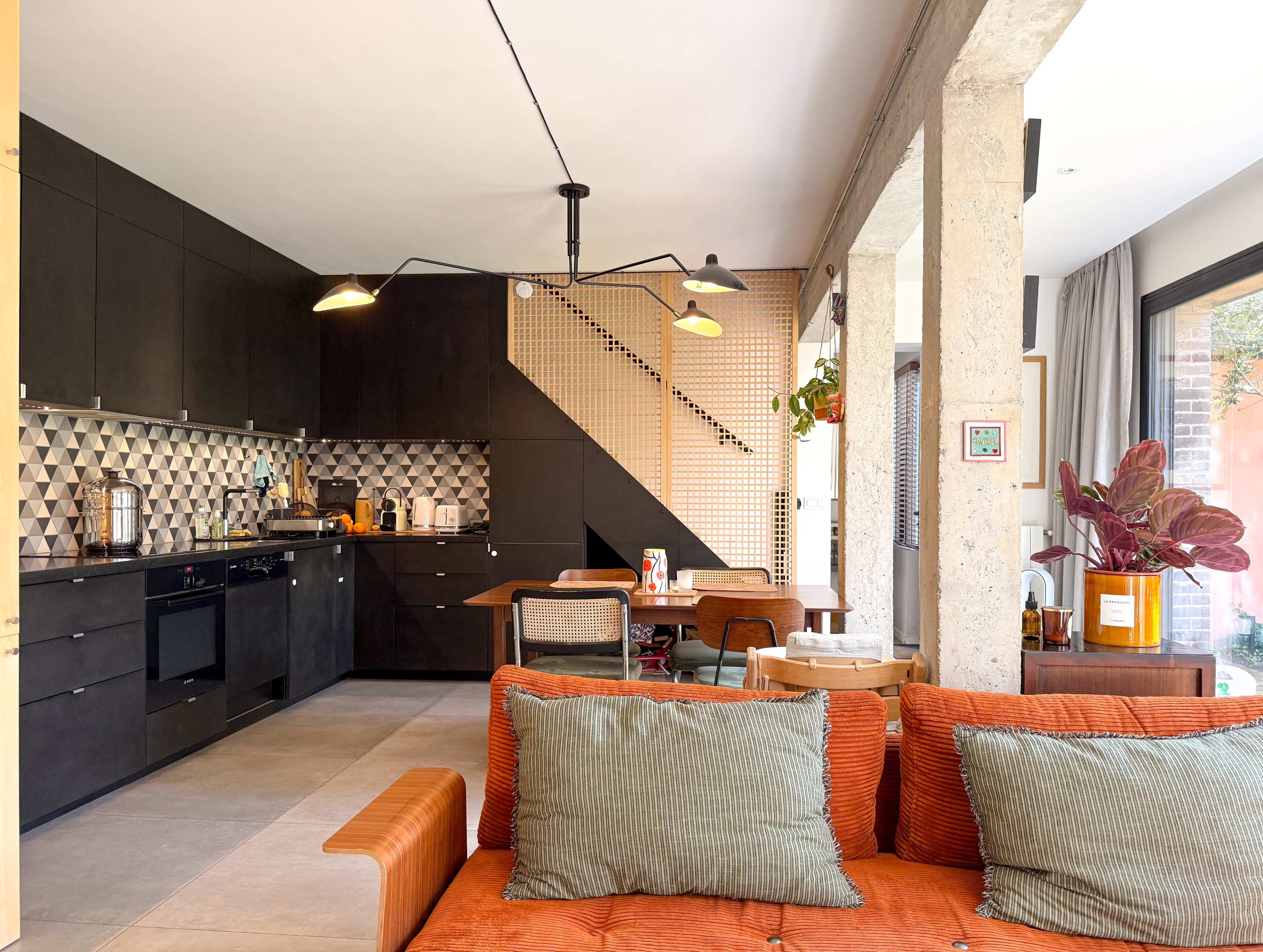 The image shows a modern kitchen and dining area featuring dark cabinetry, geometric tile backsplash, and a decorative staircase, with a cozy orange sofa in the foreground.